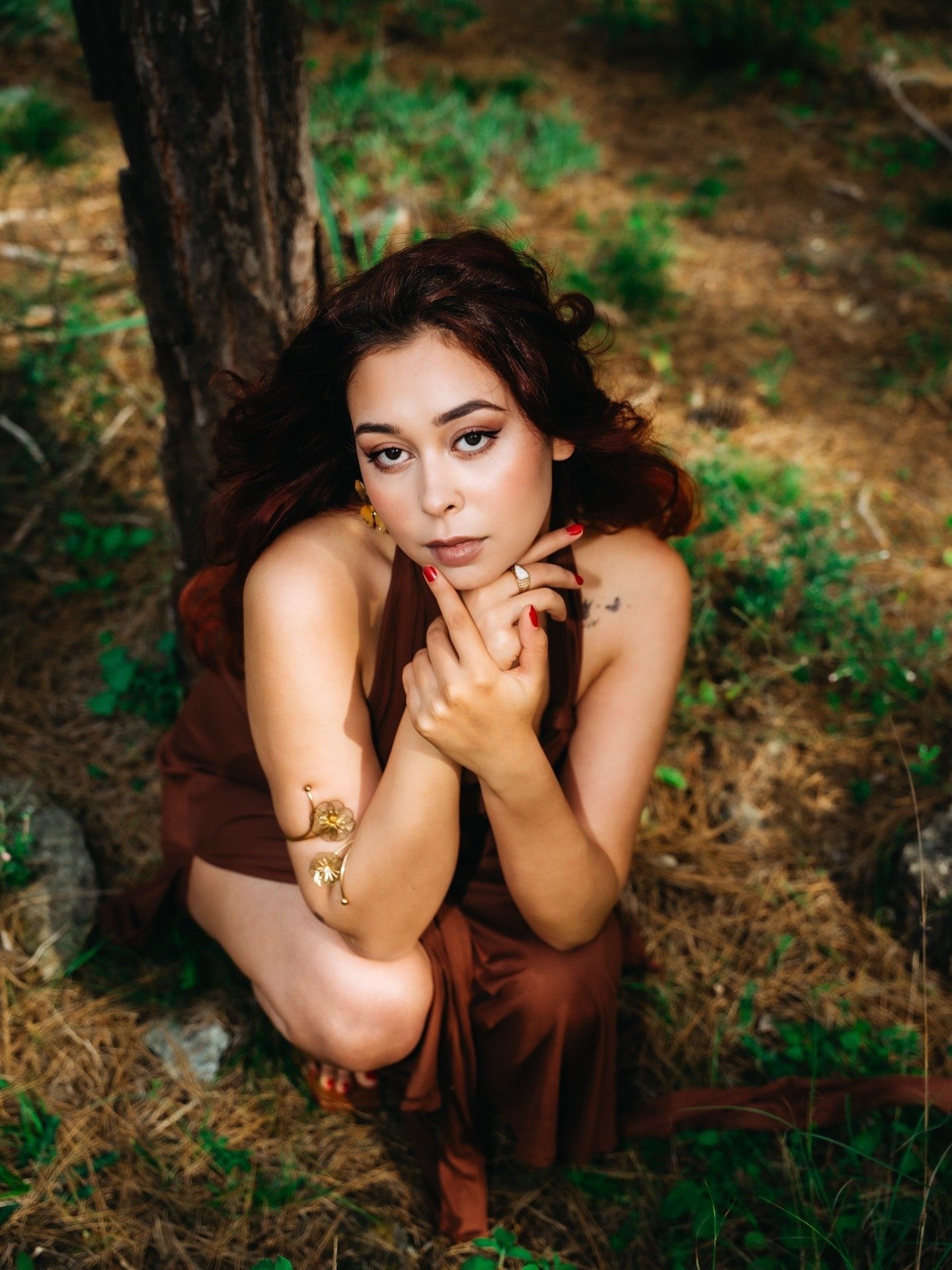 Woman in brown dress crouching by tree in forest, wearing gold snake bracelet, with flowing auburn hair.