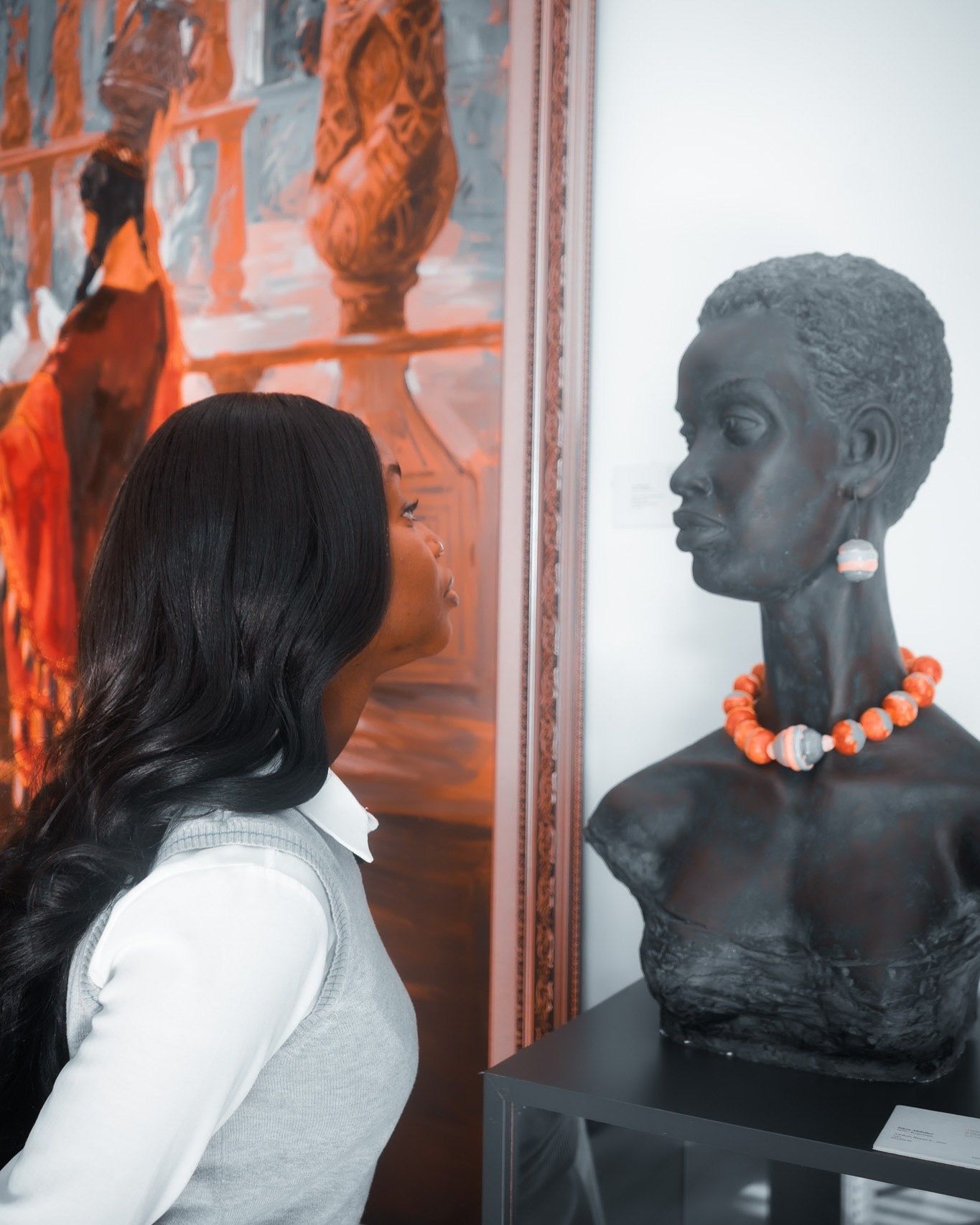 Woman gazes at a dark sculptural bust wearing an orange beaded necklace, displayed beside a vibrant orange painting in a gallery.