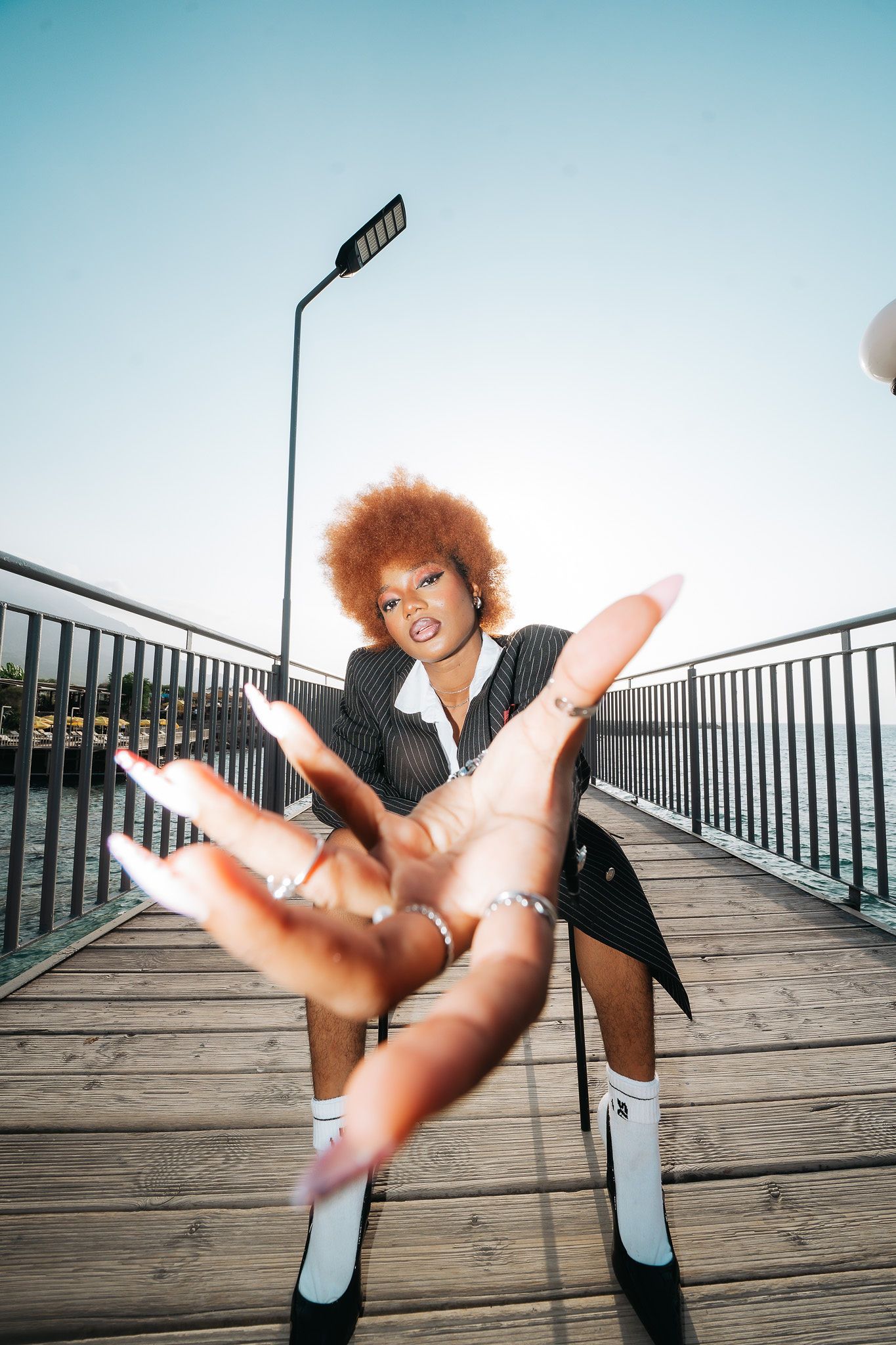 Woman with red afro in pinstripe blazer reaches toward camera on sunny wooden pier.