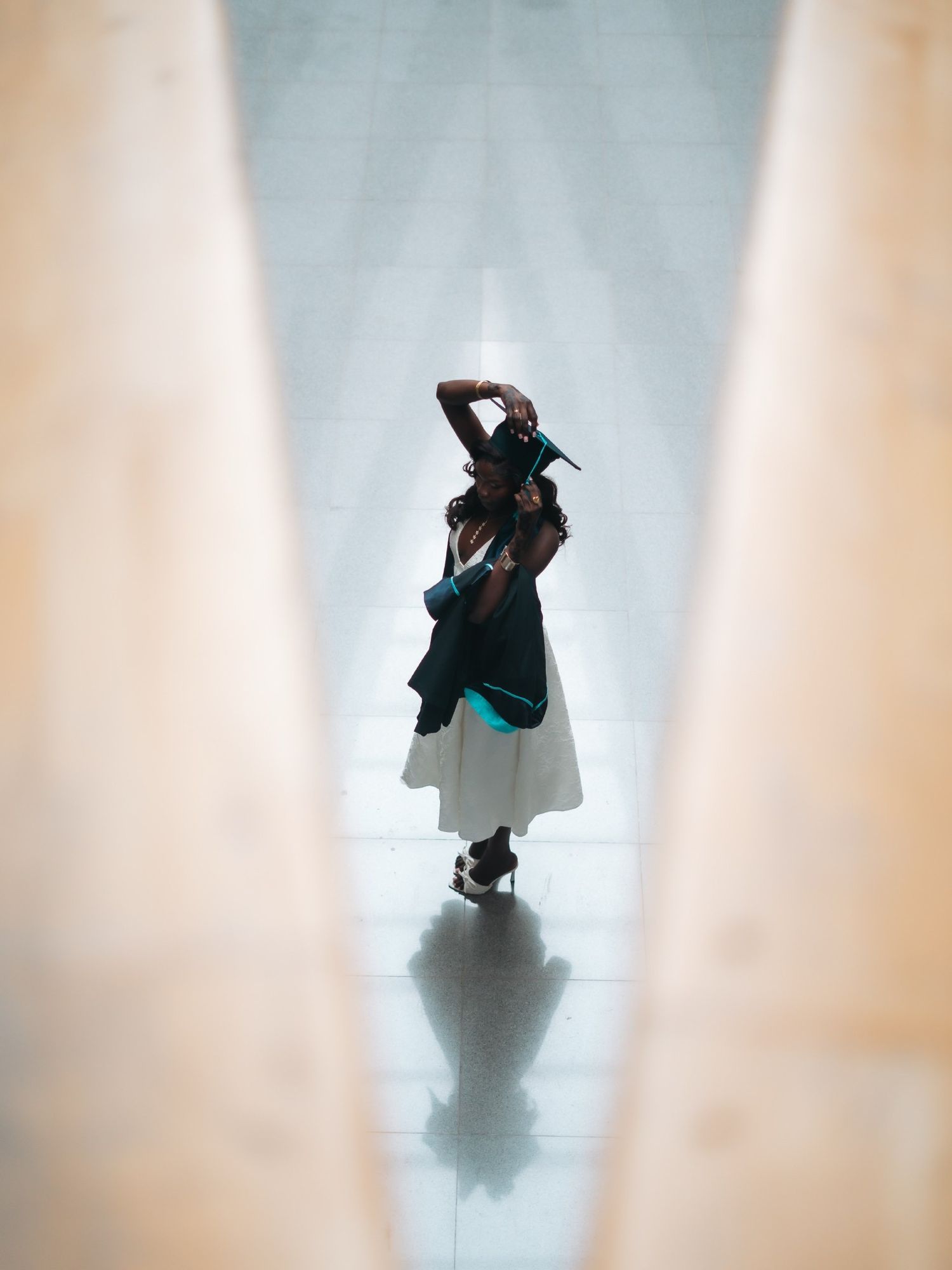 Graduate in white dress and cap poses dramatically in a bright architectural corridor.