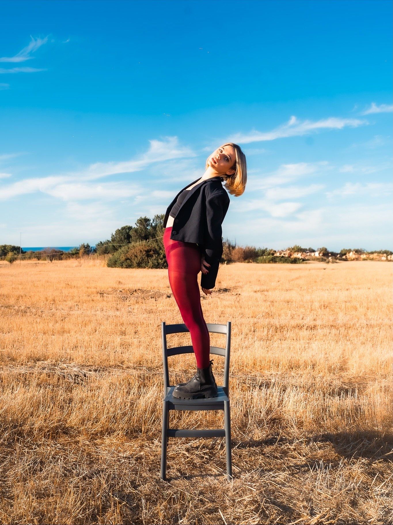 Blonde woman standing on a chair in a golden field wearing red leggings and black jacket under a blue sky.