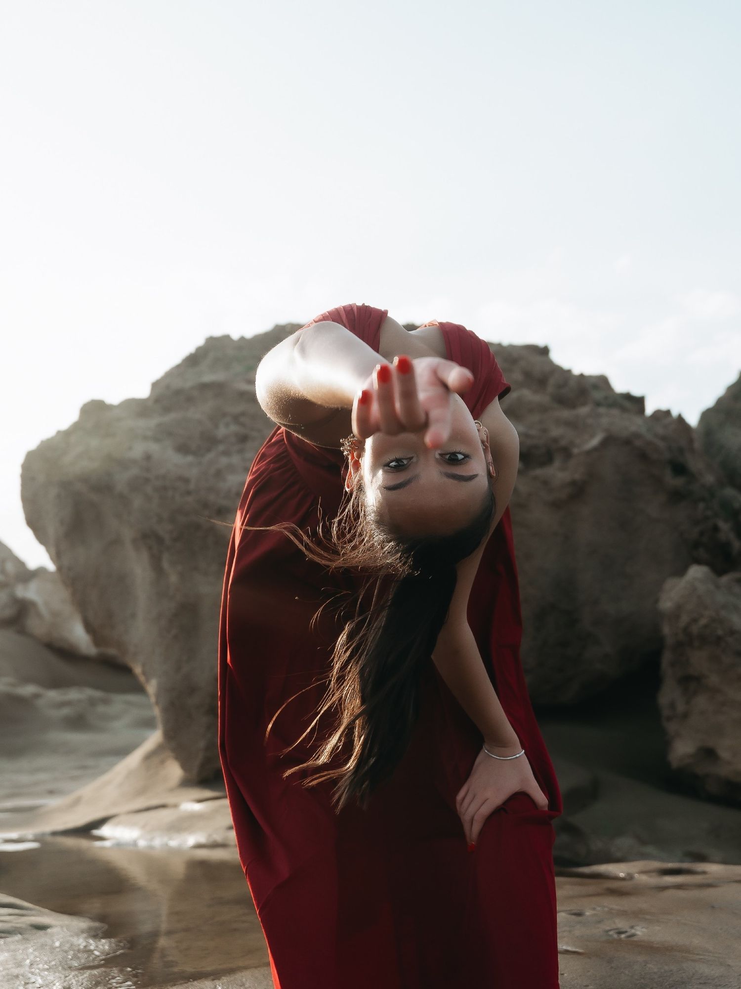 Woman in red dress reaching toward camera at rocky beach, hair flowing, with dramatic sky background.