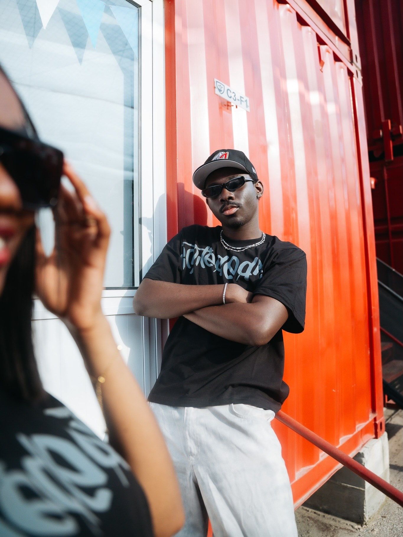 Young Black man in black cap, sunglasses, and graphic tee poses arms crossed against a red shipping container.