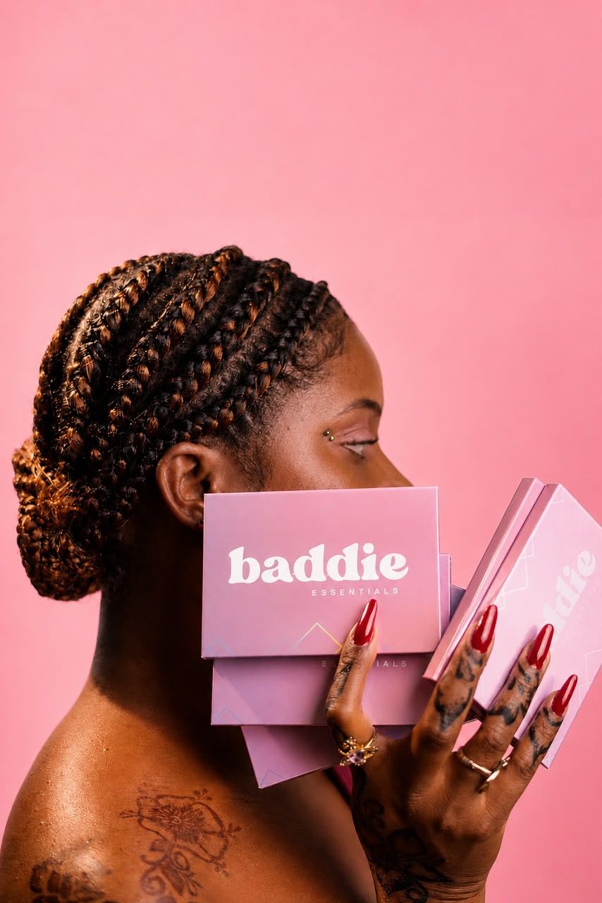 Woman with braids holding pink Baddie Experience boxes against a pink background with red nails and tattoos.