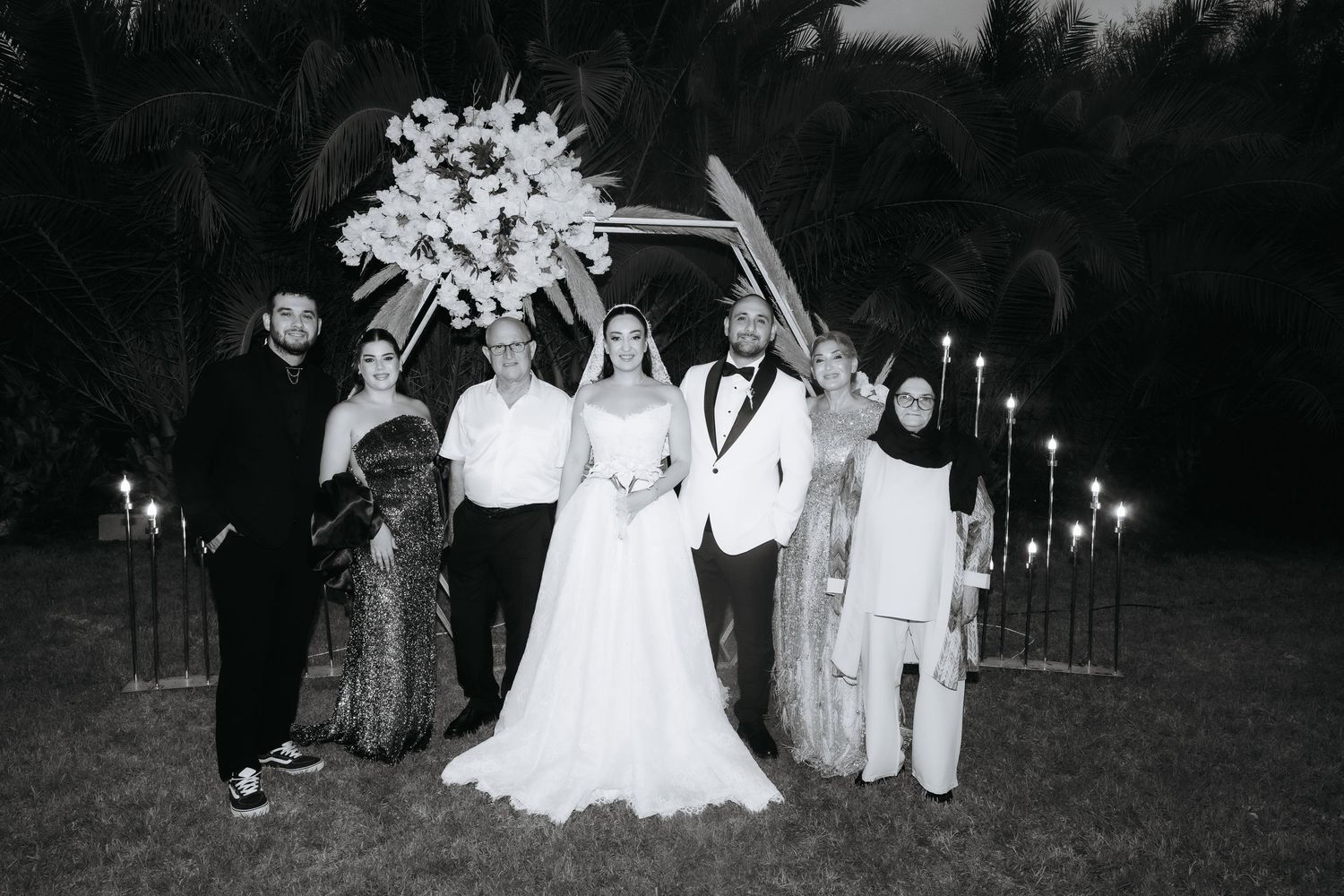 Black and white wedding photo of bride, groom, and family posing under floral arch with candles at outdoor ceremony.