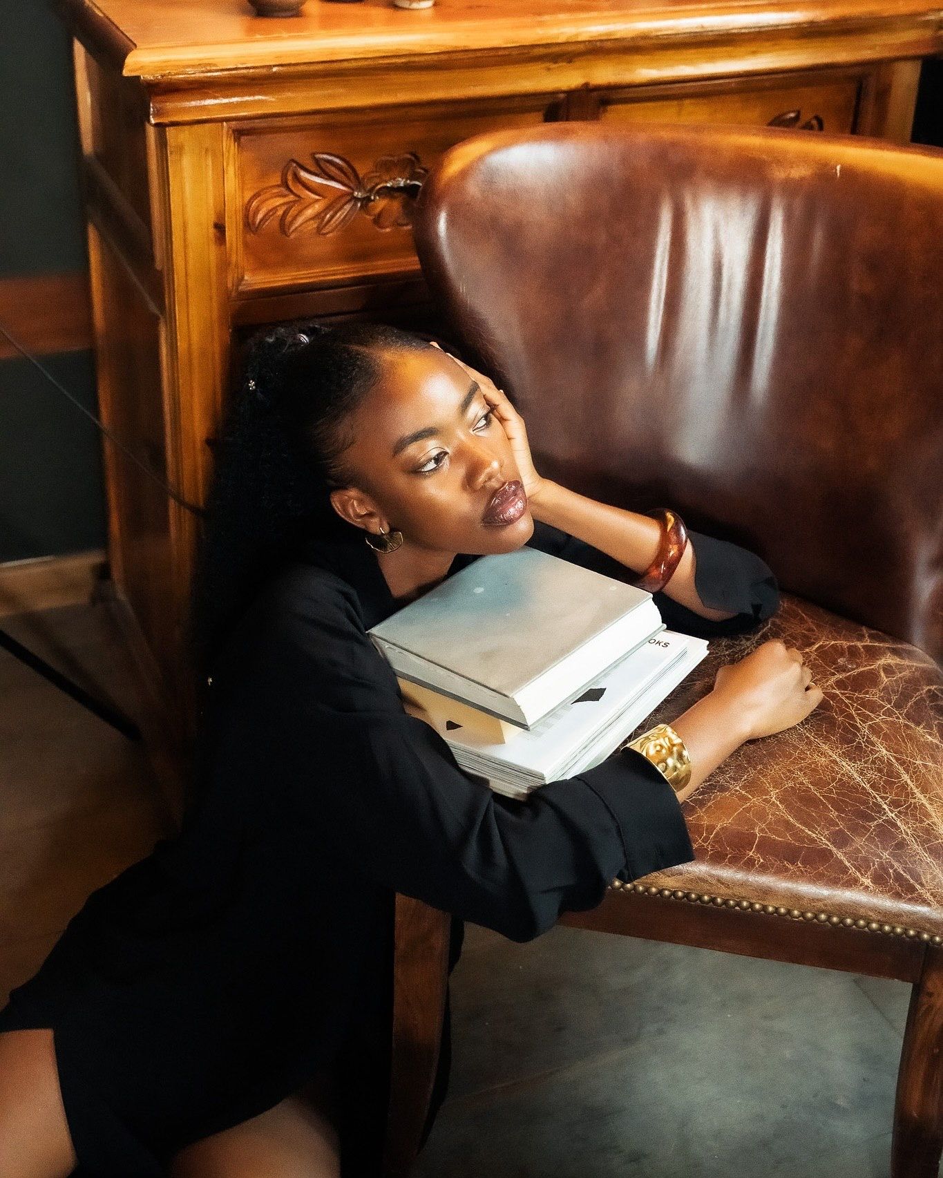 Young woman in black blazer resting on books beside a leather chair in a cozy library setting.