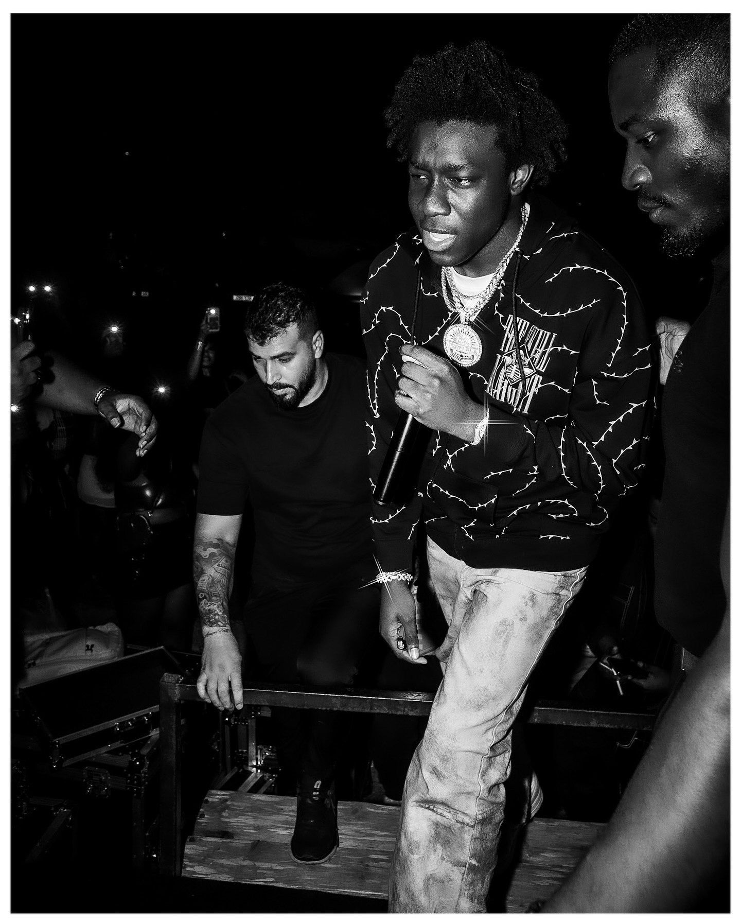 Black and white photo of a young man in a patterned jacket and chain necklace at a nightclub event.