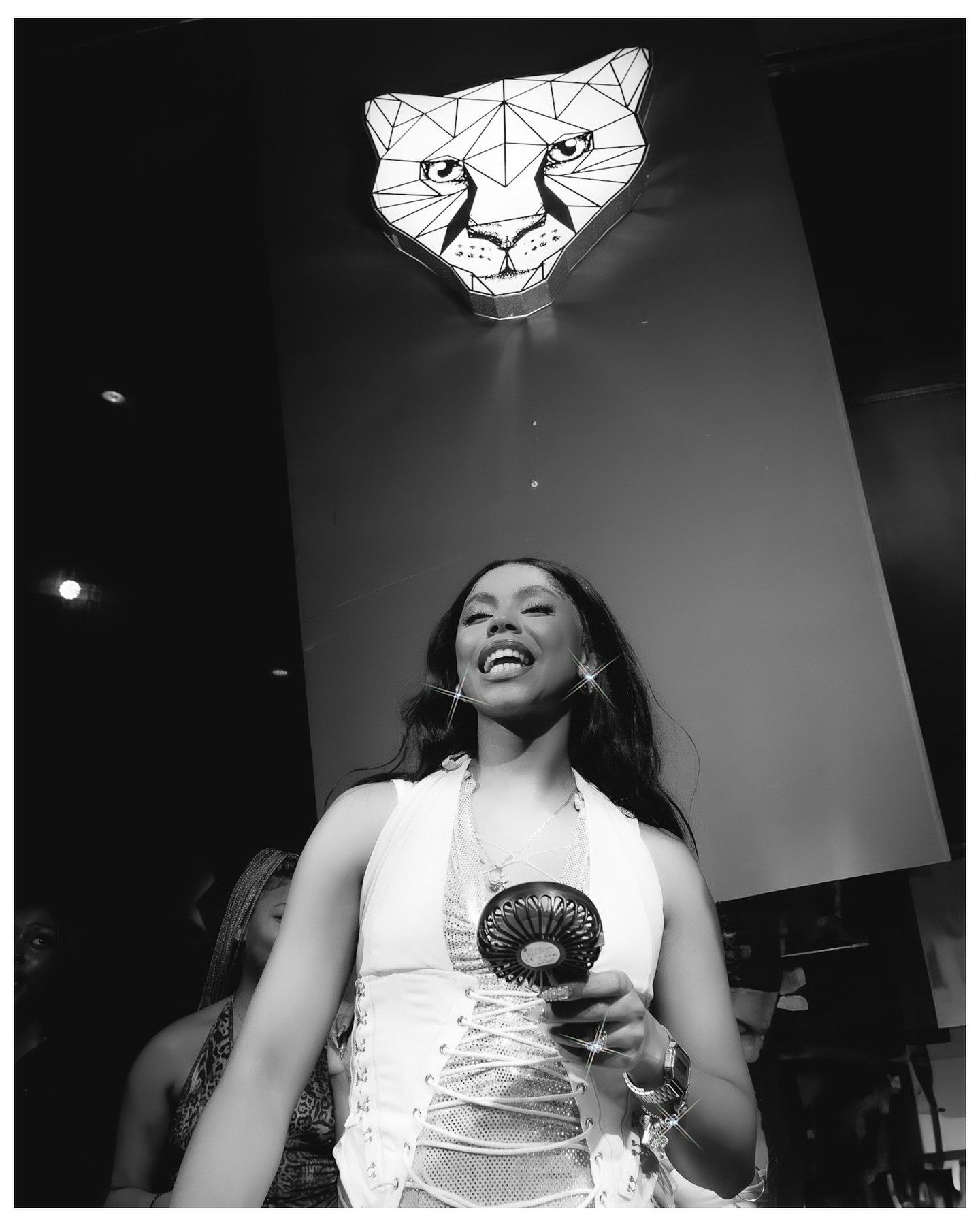 Black and white photo of a smiling woman in a white halter dress holding a fan, with a geometric cheetah neon sign above.