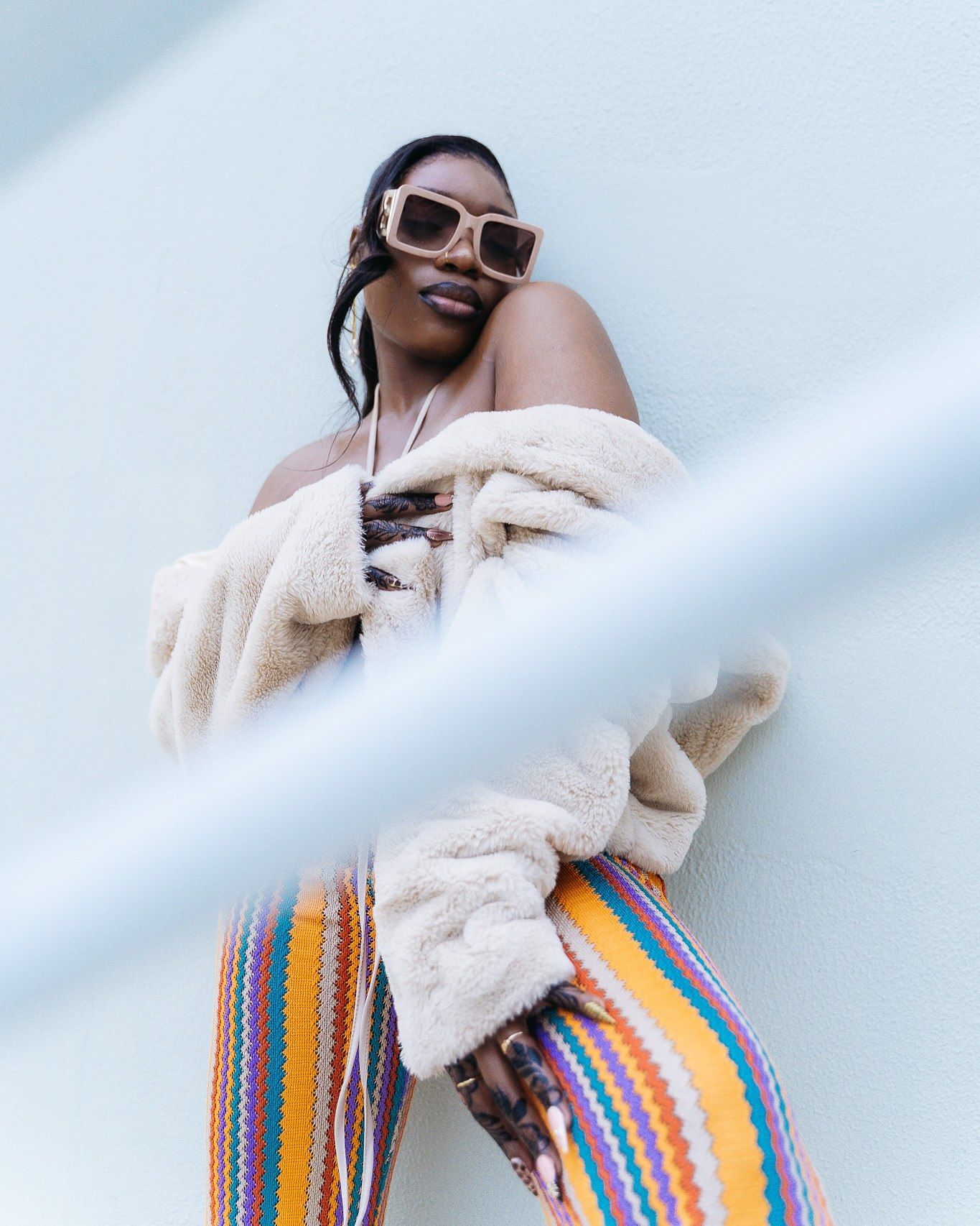 Stylish woman in colorful striped pants and cream fur jacket poses against a white wall wearing sunglasses.