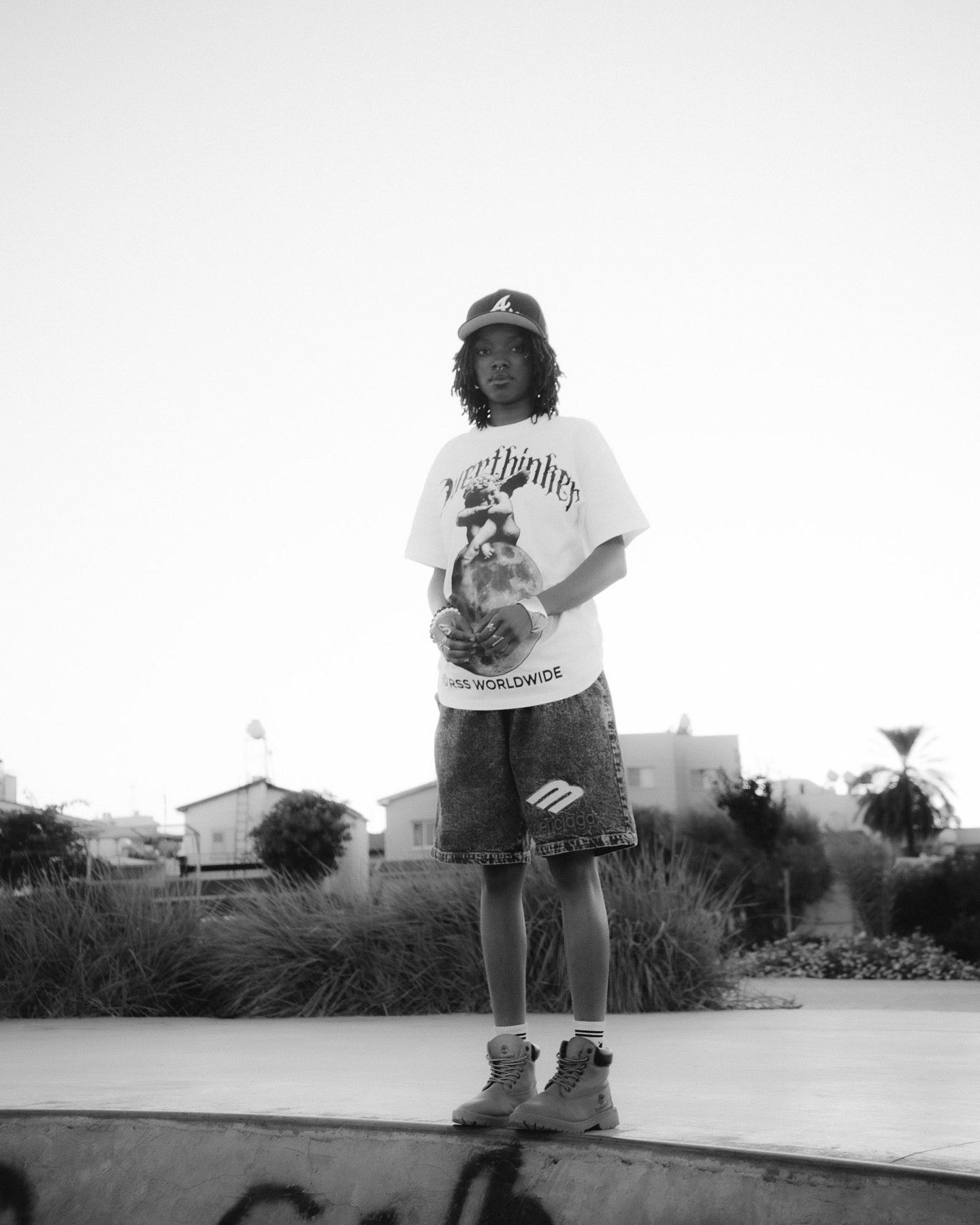 Young skater in white graphic tee, denim shorts, and cap poses at a skate park in black and white photo.