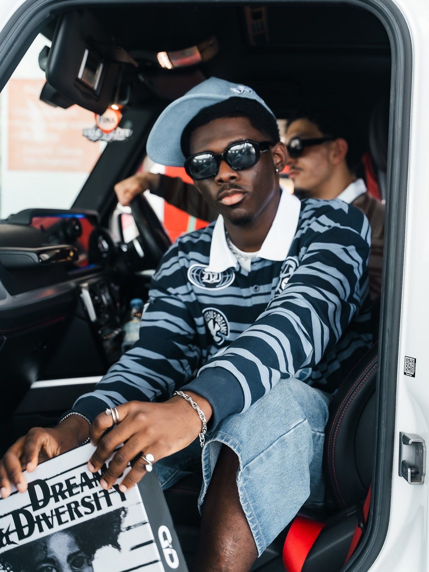 Young man in striped sweater and sunglasses sits in white car holding Dream & Diversity sign with friend behind him.