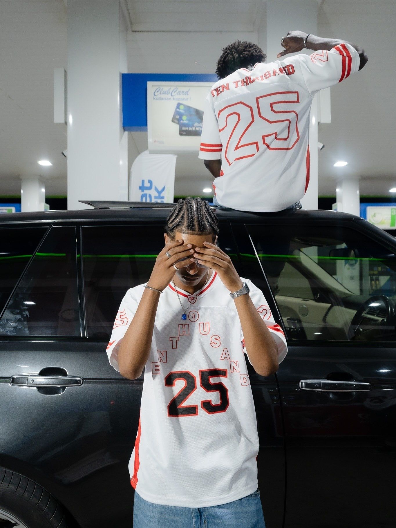 Two people wearing matching white number 25 jerseys pose near a black SUV at a gas station.