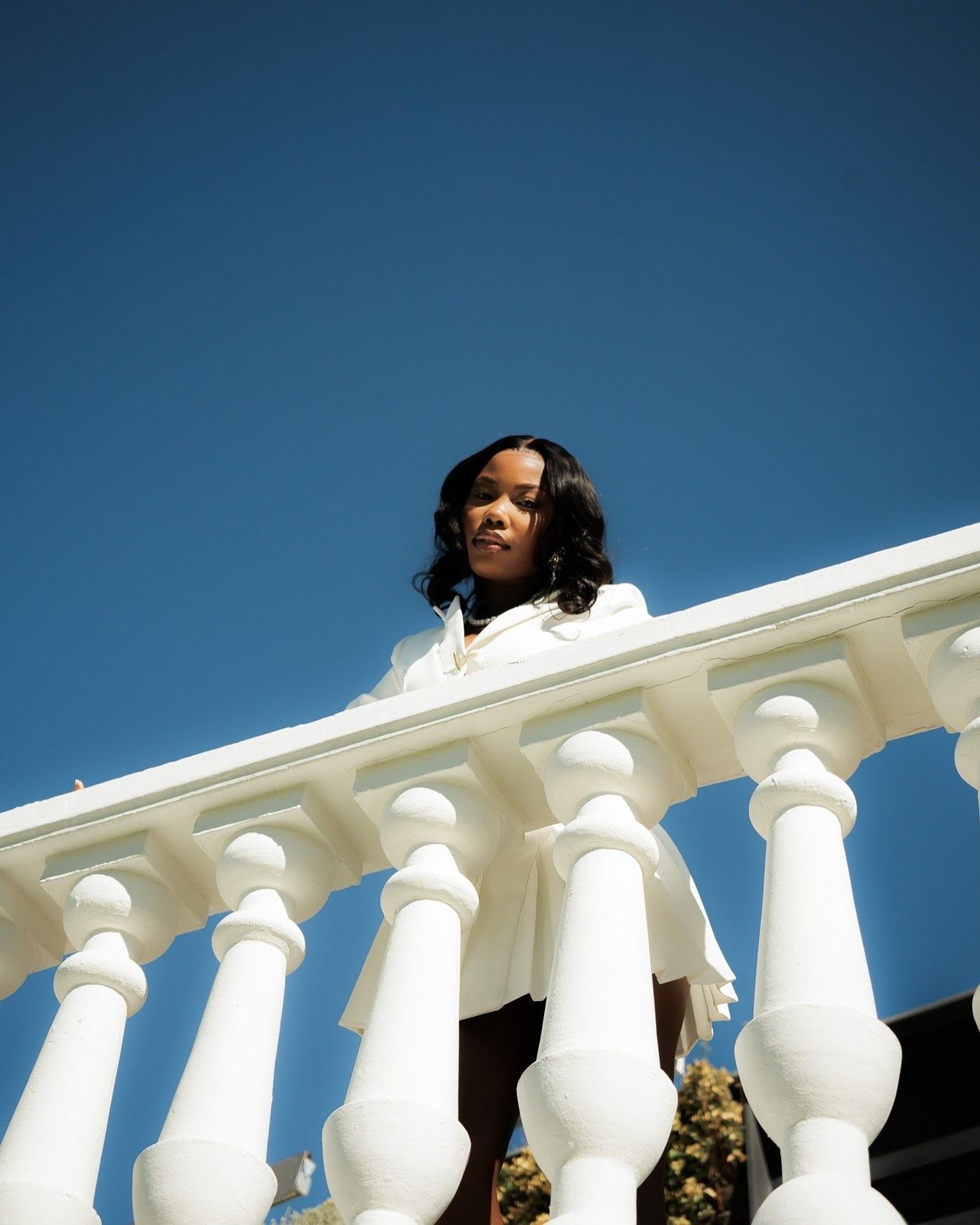 Young woman in white outfit leaning over white balustrade railing against clear blue sky.