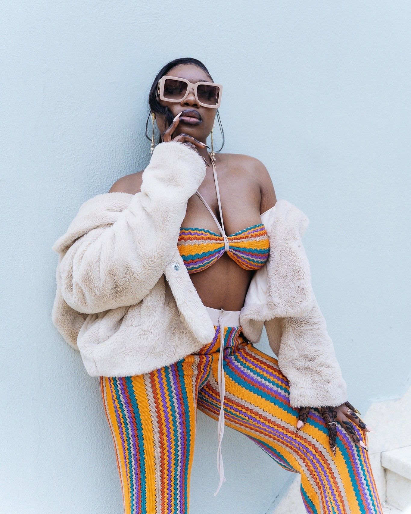 Woman in colorful striped bikini top and pants with cream faux fur jacket and square sunglasses posing against light blue wall.