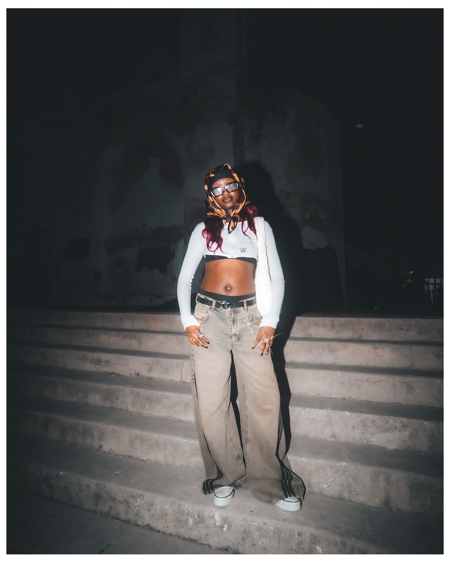 Person in white crop top and baggy tan jeans posing on outdoor steps at night with colorful hair accessories.