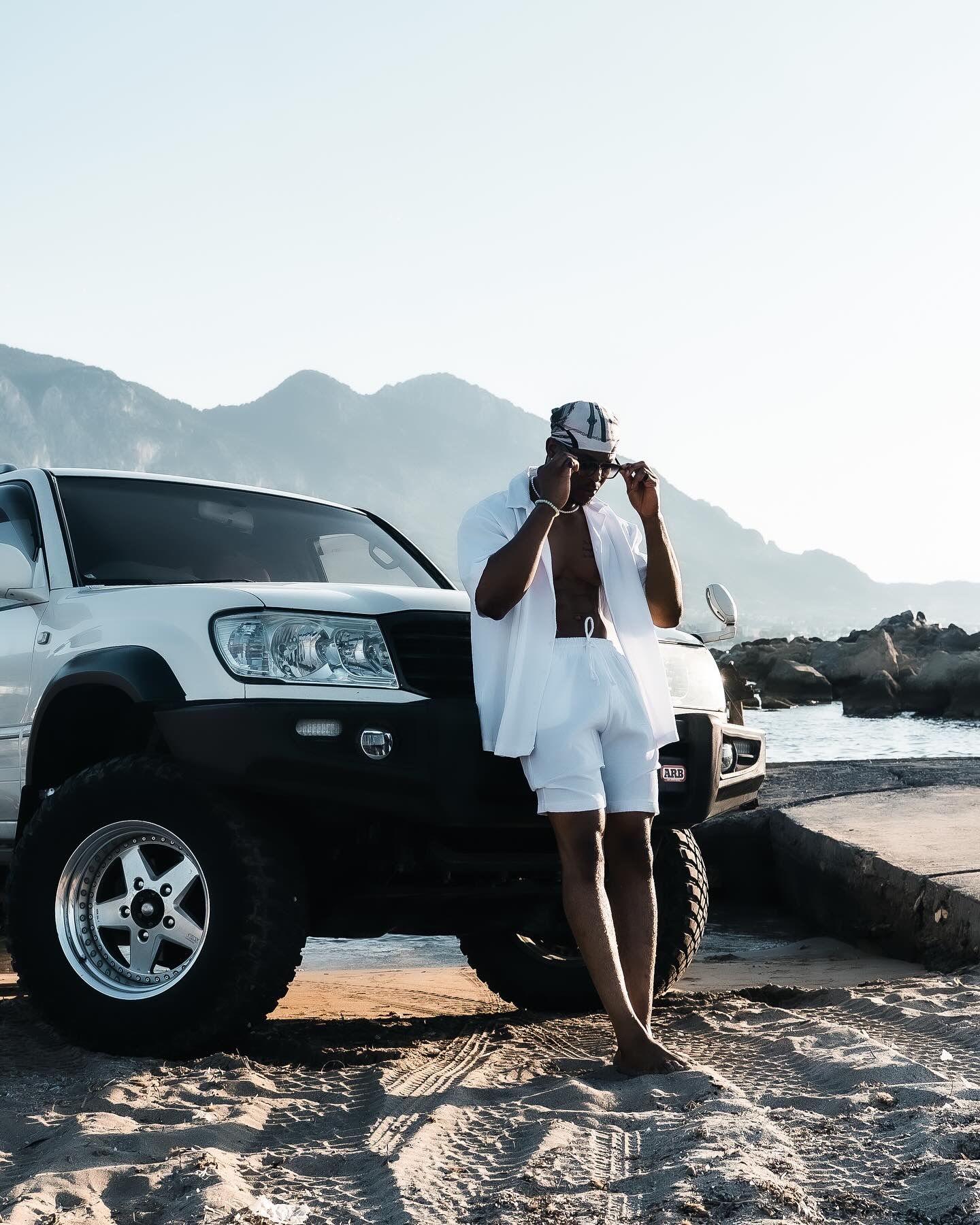 Man in white outfit standing beside a white Toyota SUV on a sandy beach with mountains in the background.