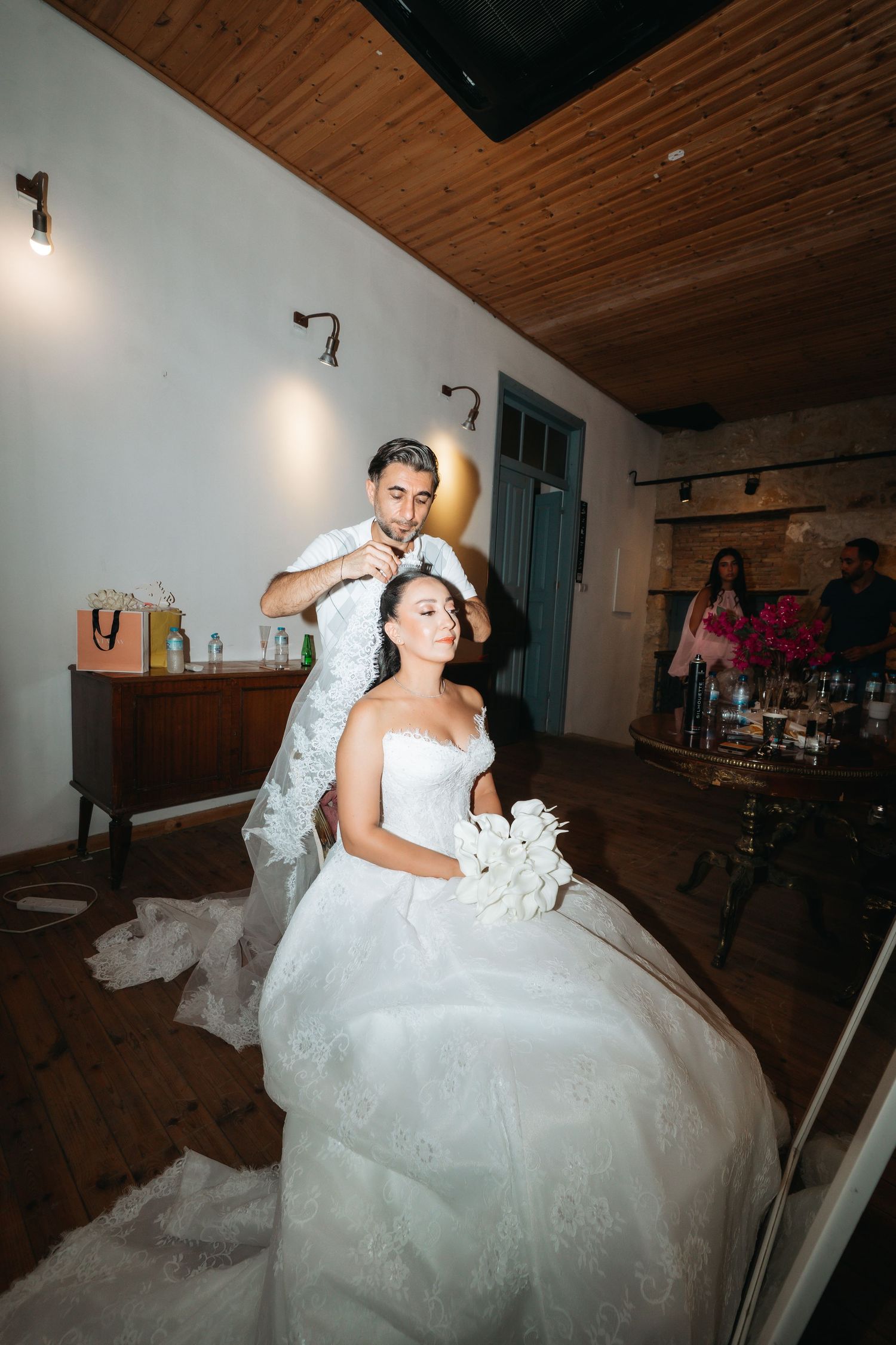 Groom placing veil on seated bride in white gown holding bouquet during indoor wedding ceremony.