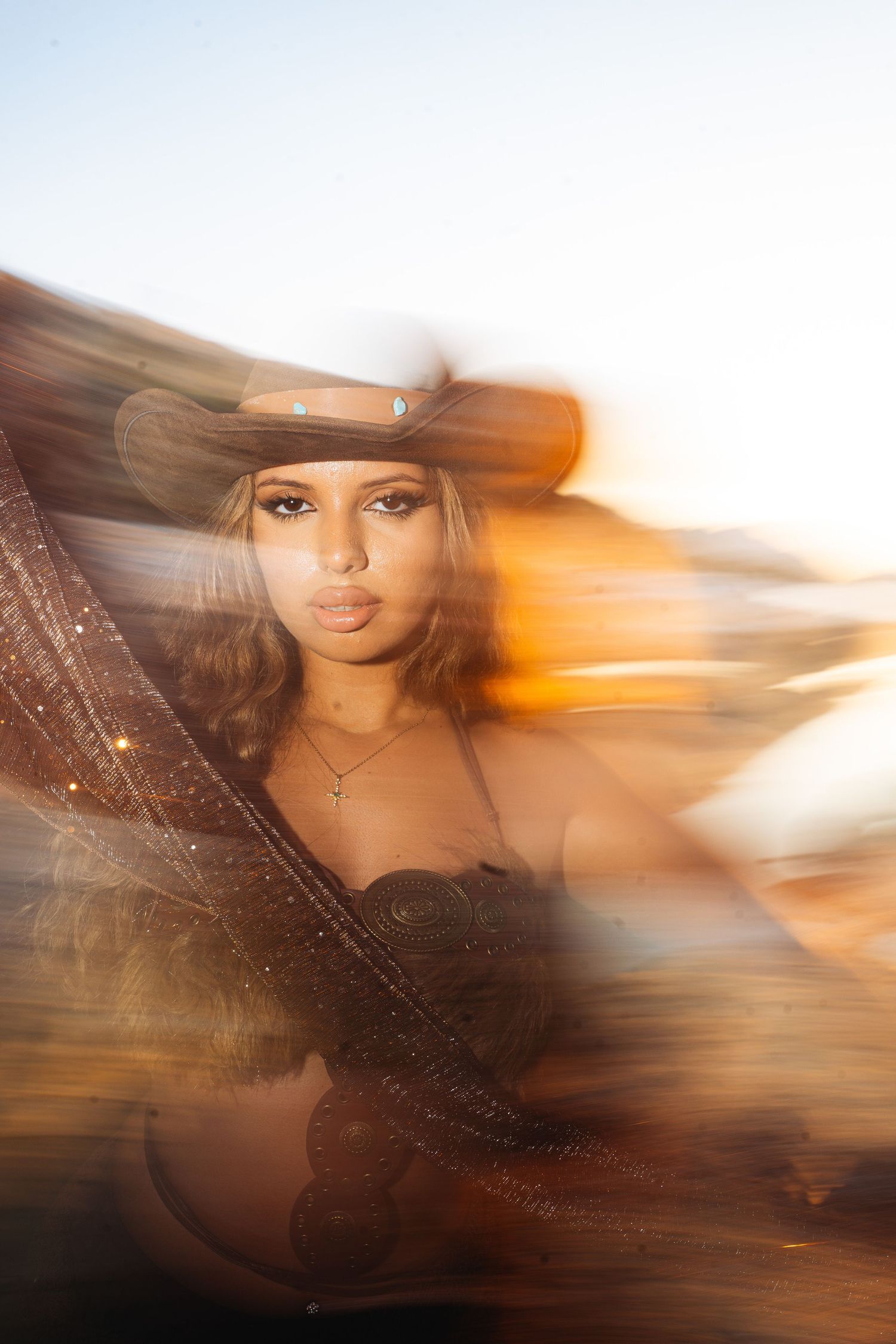 A young woman in a cowboy hat poses dramatically with flowing fabric in a warm, blurred golden-hour outdoor setting.