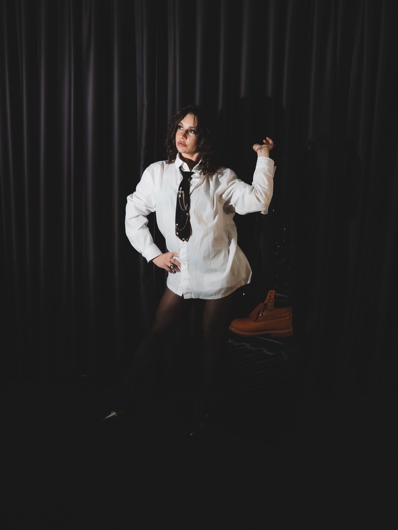 Woman in white shirt black tie and tights poses standing beside orange boots in dark studio.