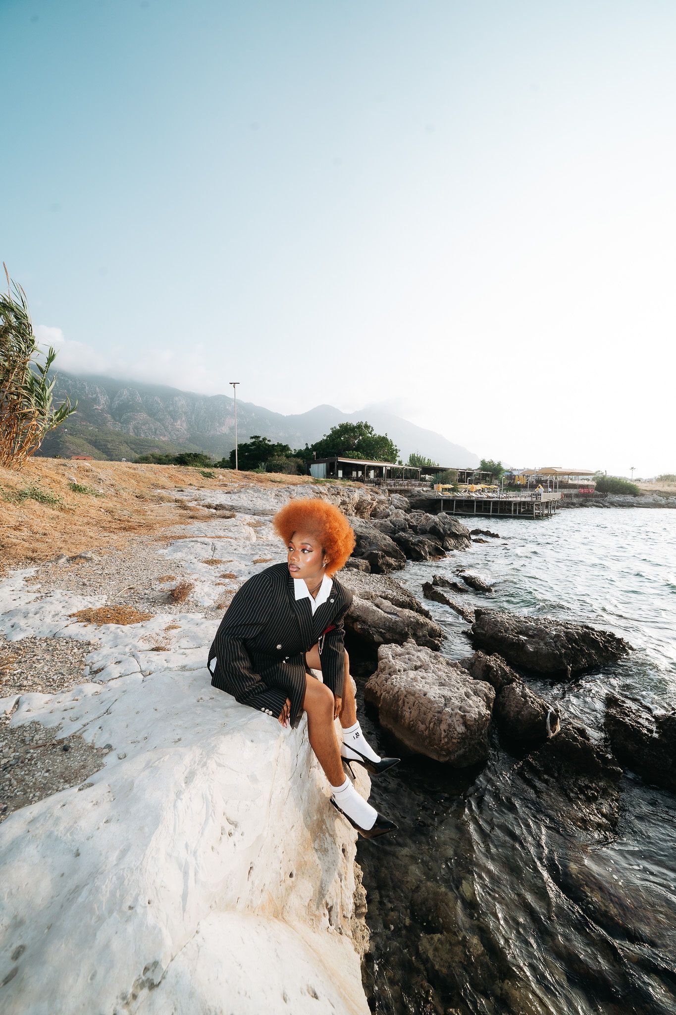 Woman with red afro in black outfit sitting on white rocks by a scenic coastal waterfront.