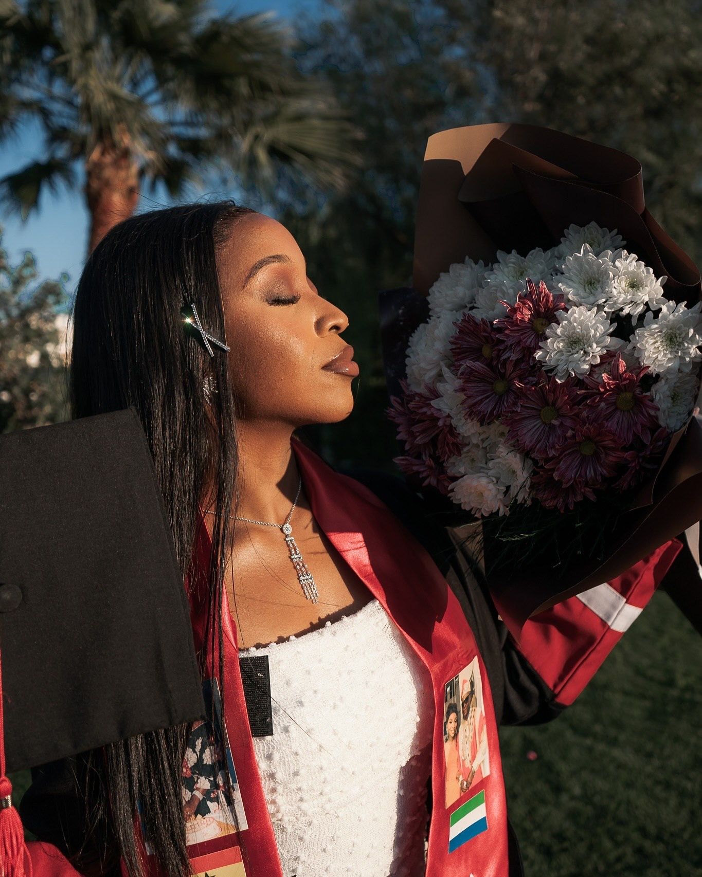 Young Black woman in maroon graduation gown holds bouquet of white and red flowers with eyes closed.