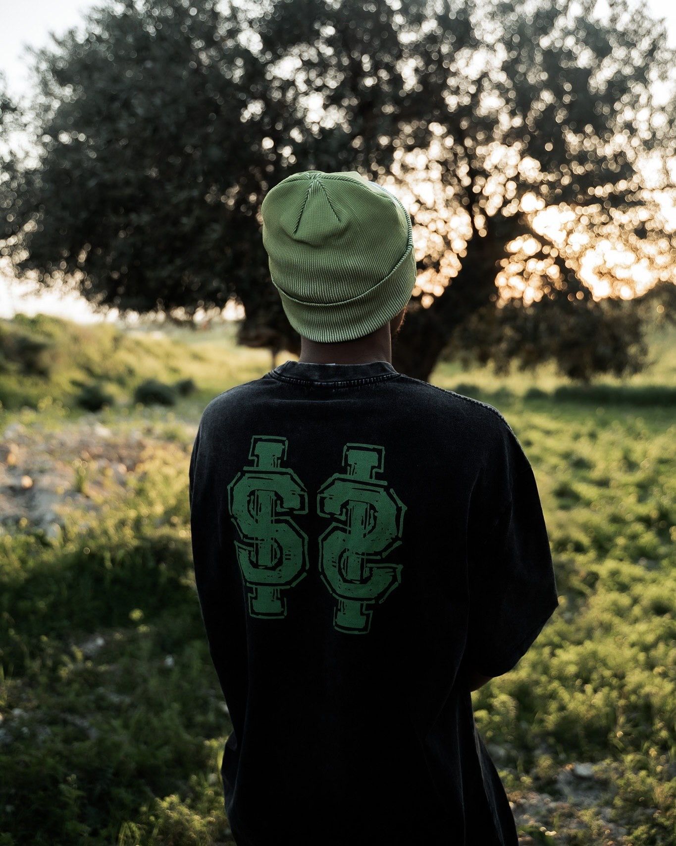 Person wearing black sweatshirt with green dollar sign logo and green beanie, standing outdoors near trees at sunset.