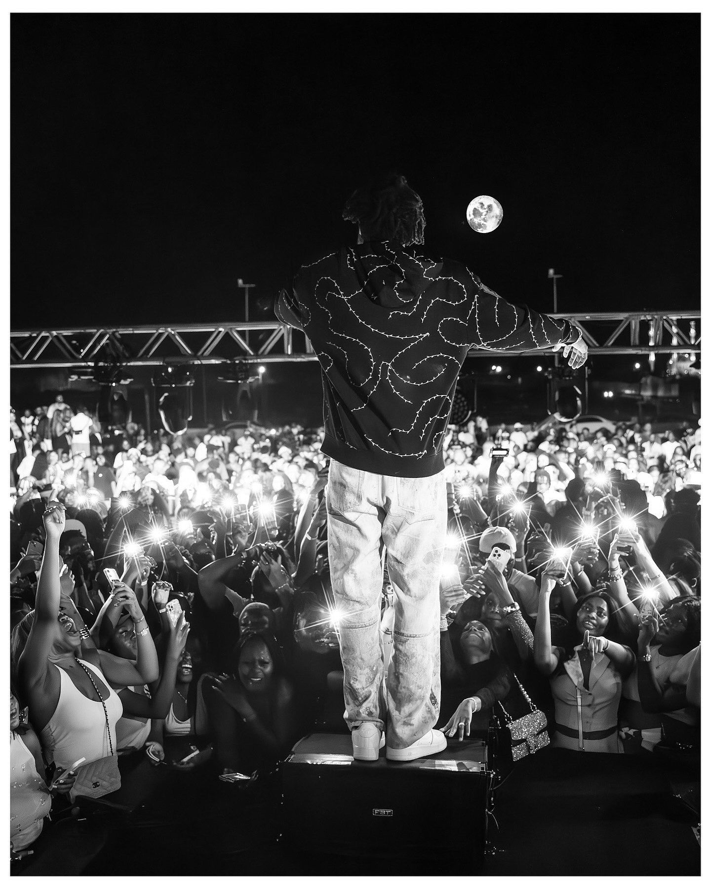 Black and white photo of a performer on stage facing a massive crowd with phone lights raised under a full moon.
