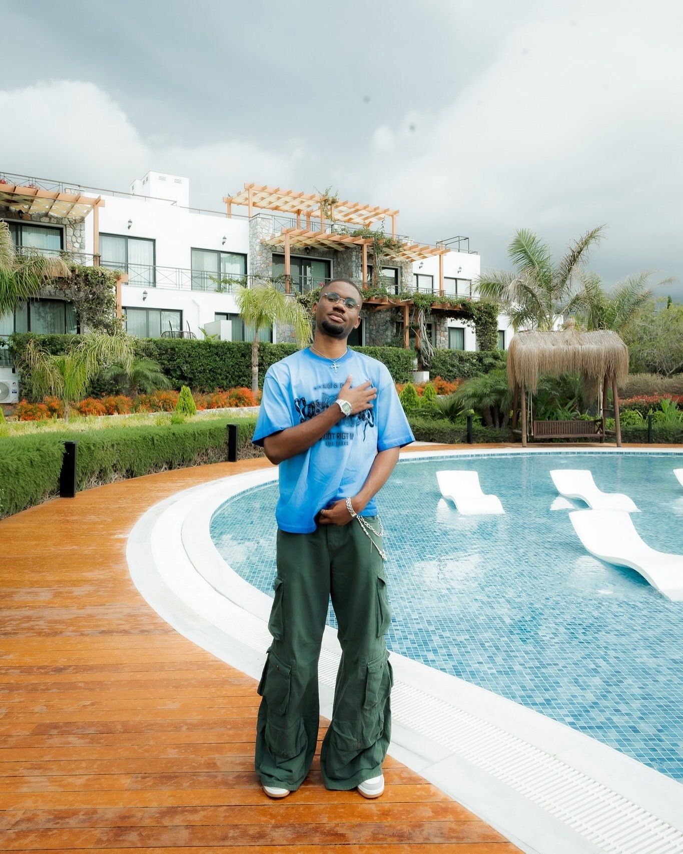 Young man in blue tee and green cargo pants posing poolside at a luxury resort with white loungers.