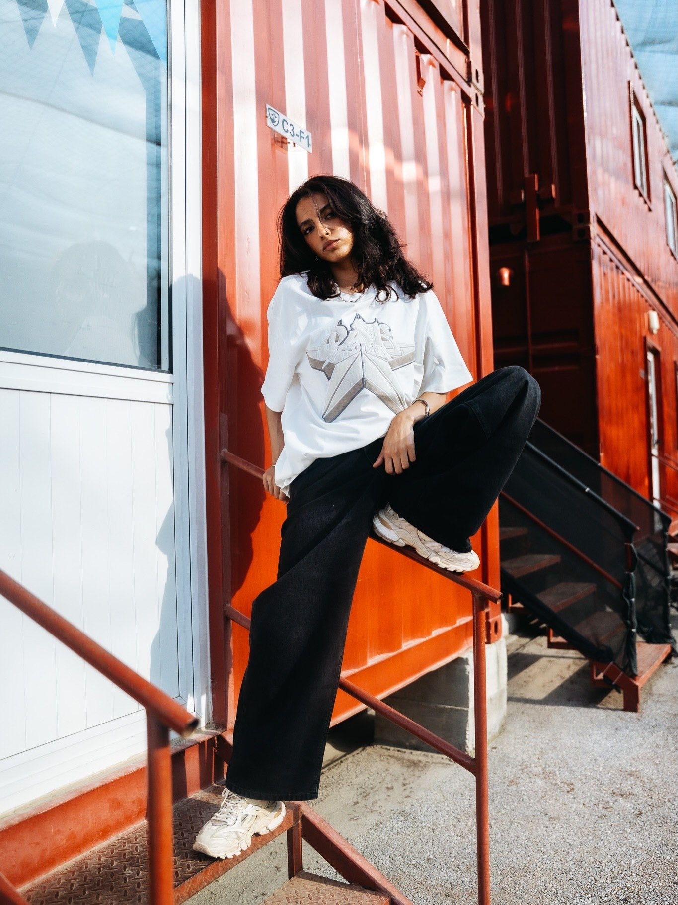 A young woman in a white graphic tee and black wide-leg pants poses on metal stairs by a red shipping container.
