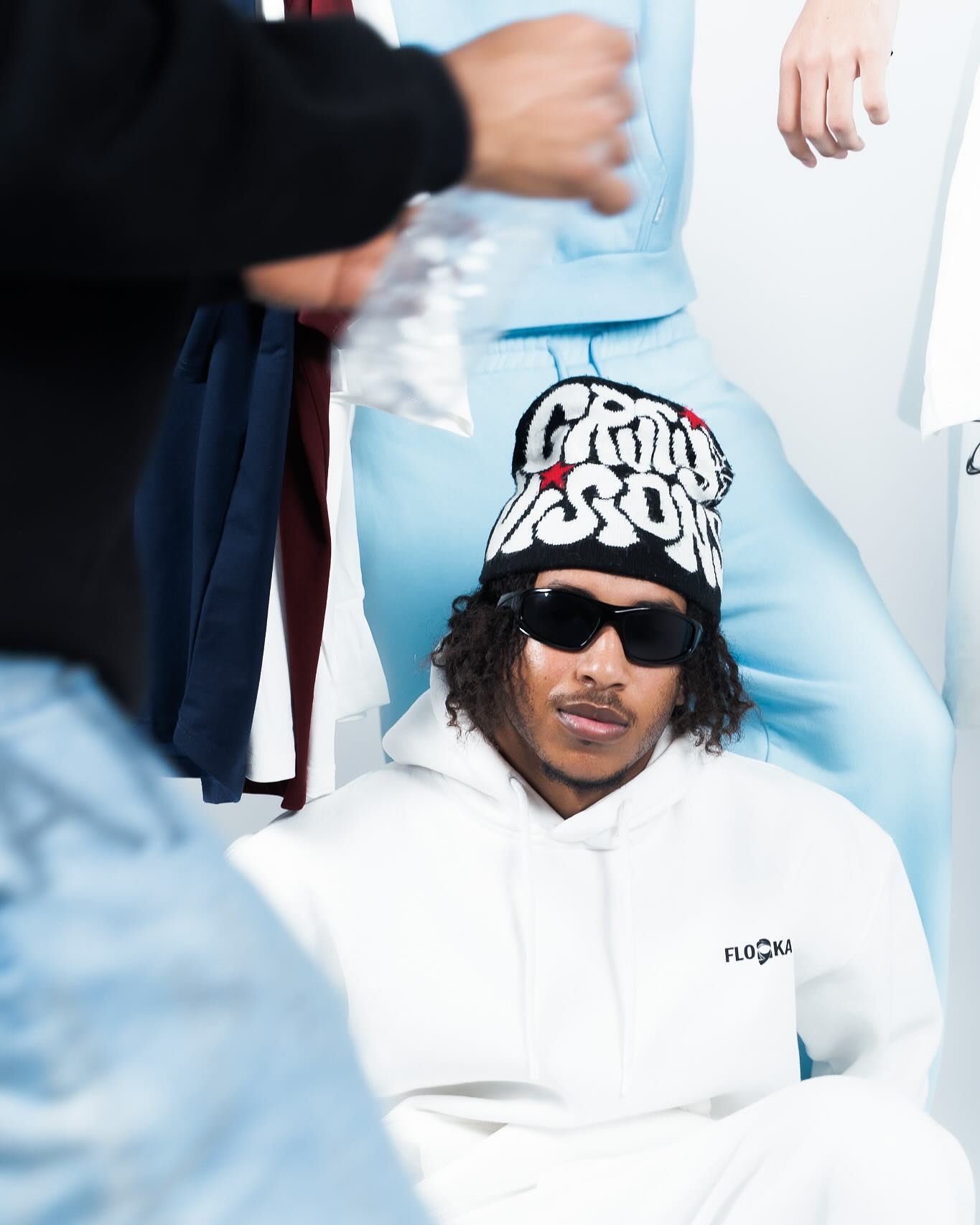 Young man in white Flo&Ka hoodie and black beanie with sunglasses sits among people in light blue streetwear.