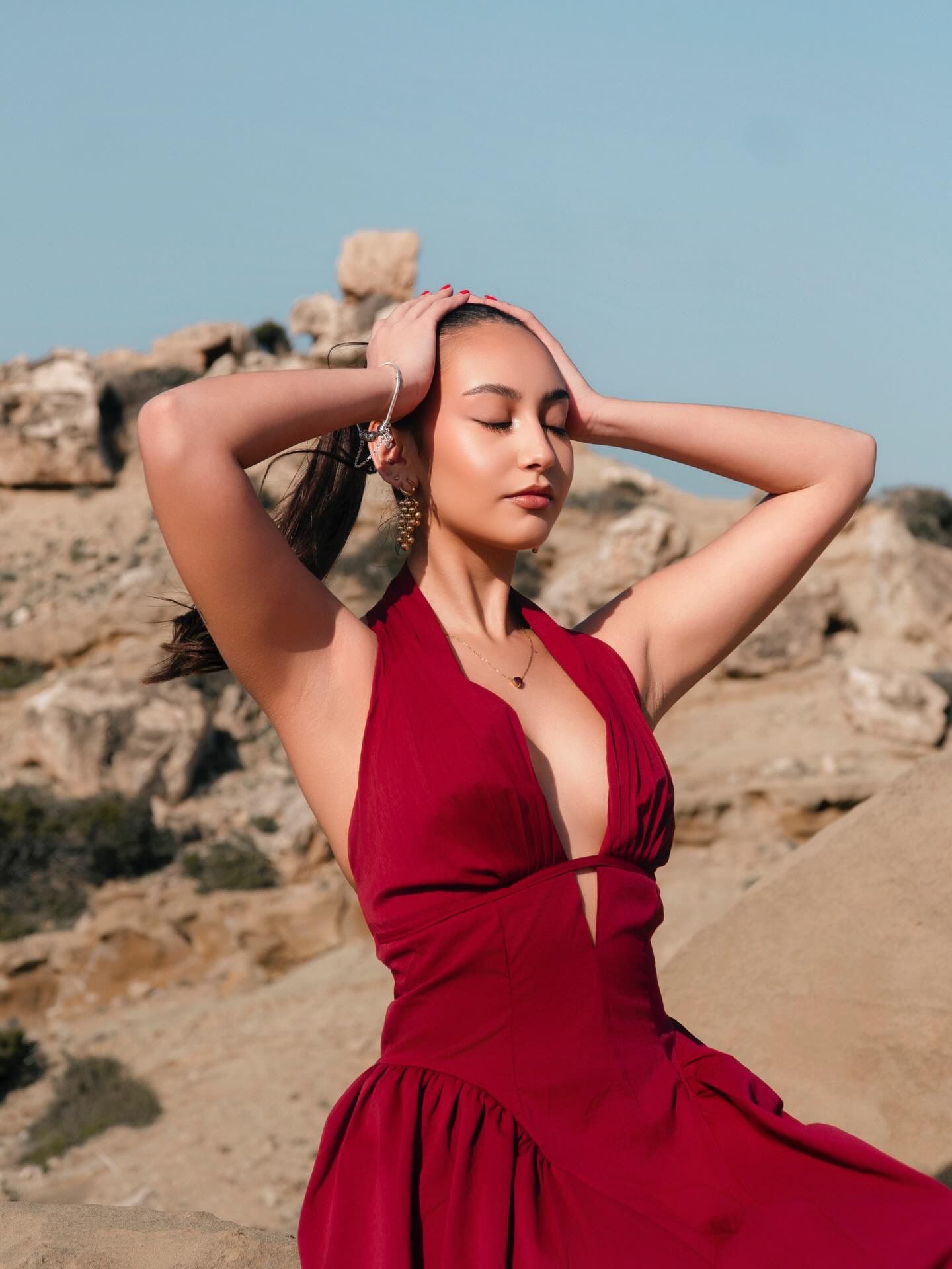 Woman in red halter dress poses gracefully on rocky terrain under blue sky.