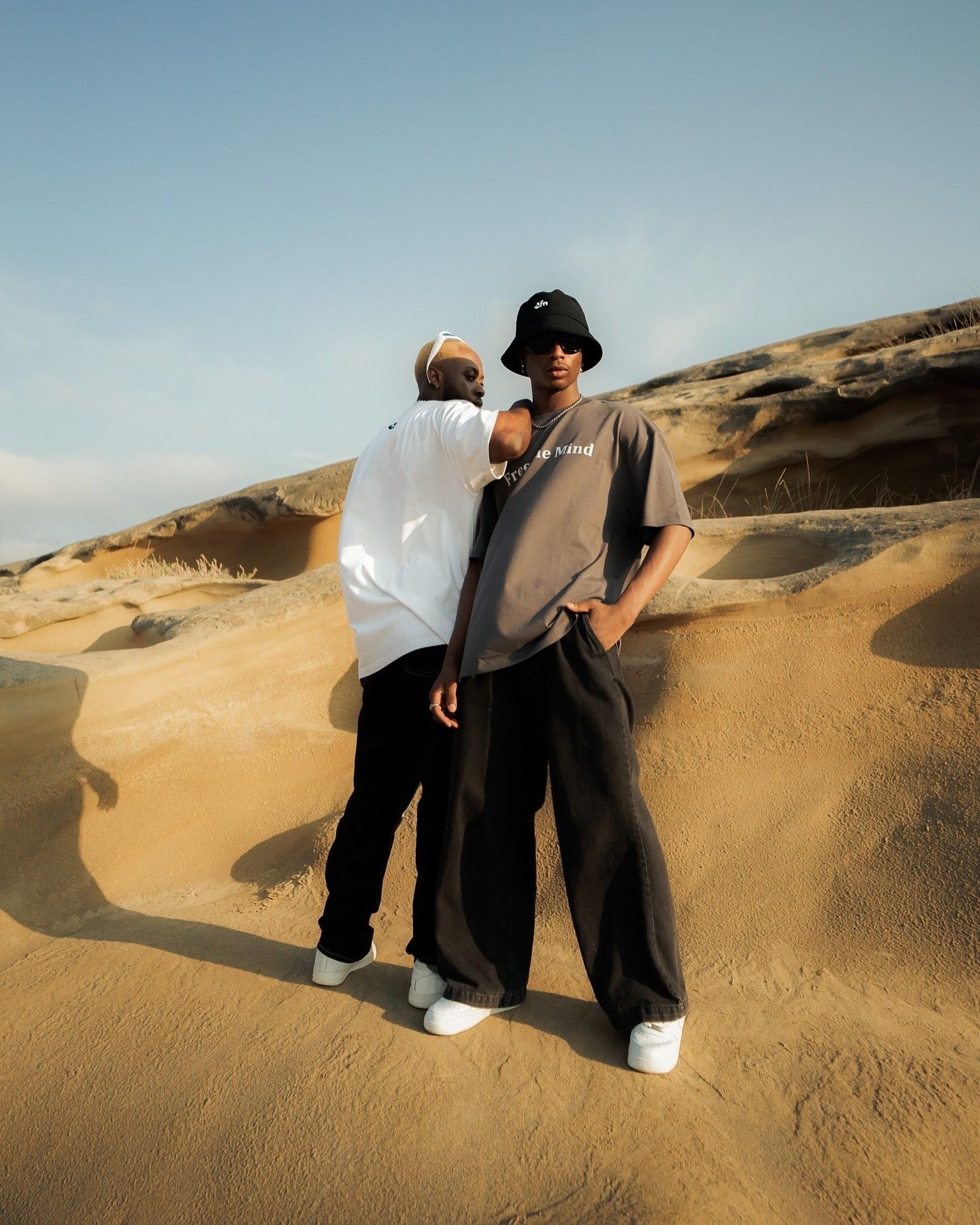 Two friends pose on golden sand dunes wearing streetwear, one in a bucket hat and wide-leg pants, desert landscape behind.
