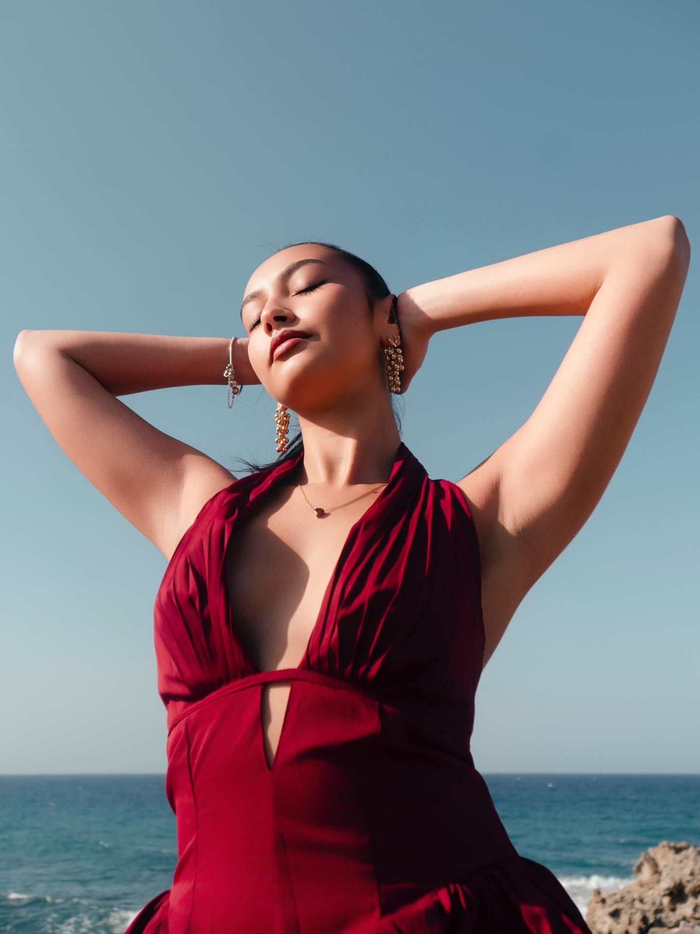 Woman in red halter dress poses with hands behind head against blue sky and ocean backdrop.