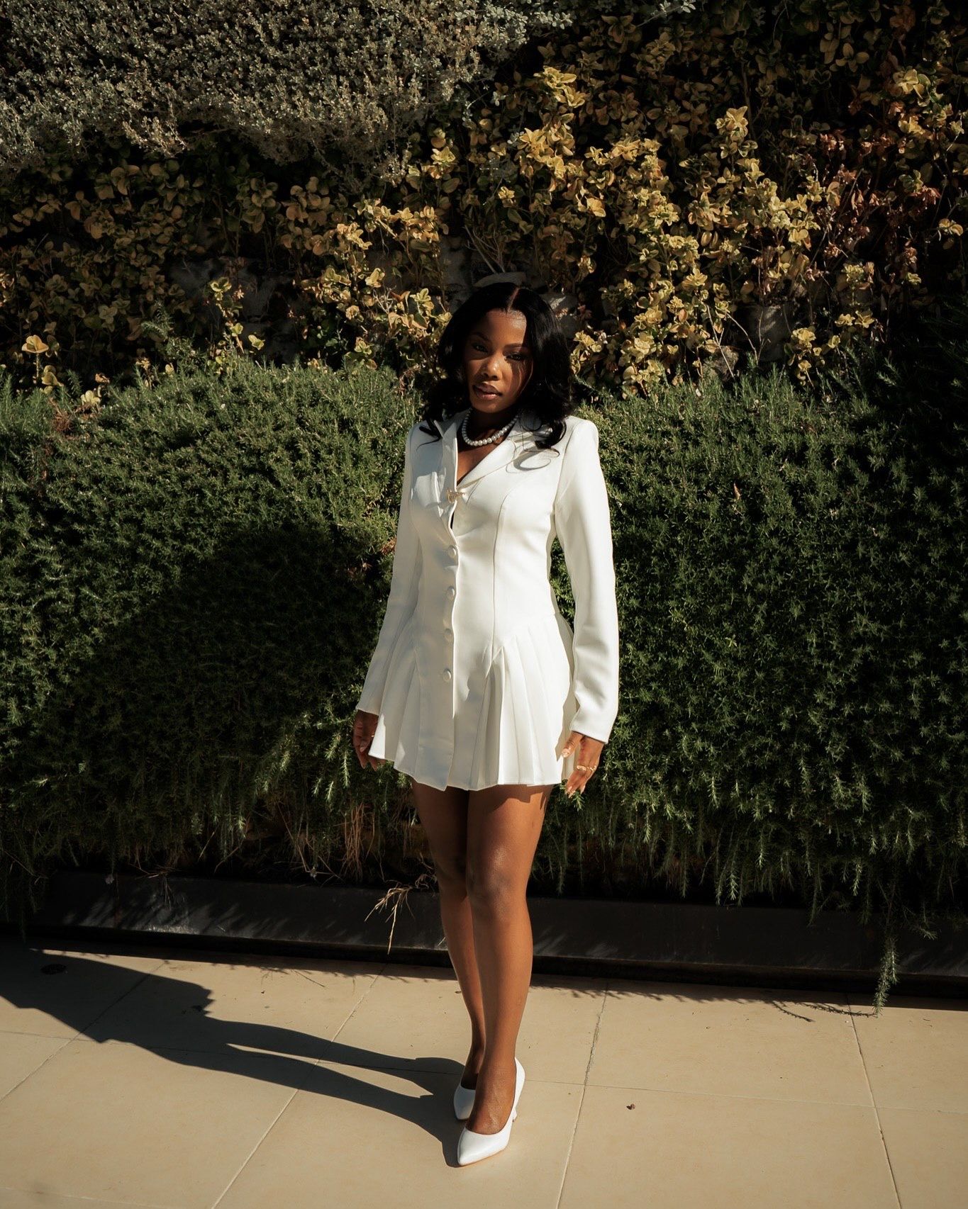 Woman in white long-sleeve mini dress and white heels posing outdoors against green hedge wall.