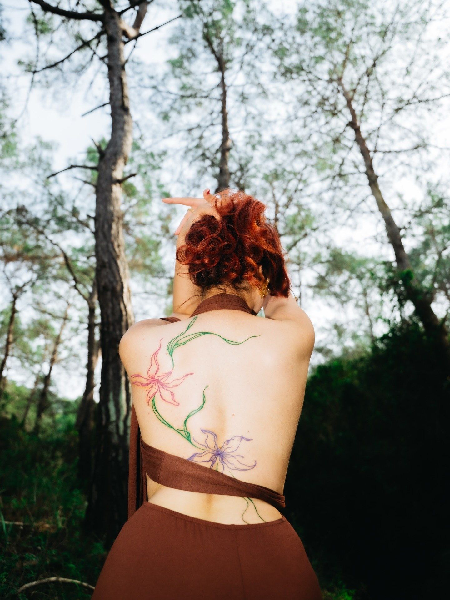 Woman with red hair and floral back tattoo standing in a forest wearing a brown halter top.