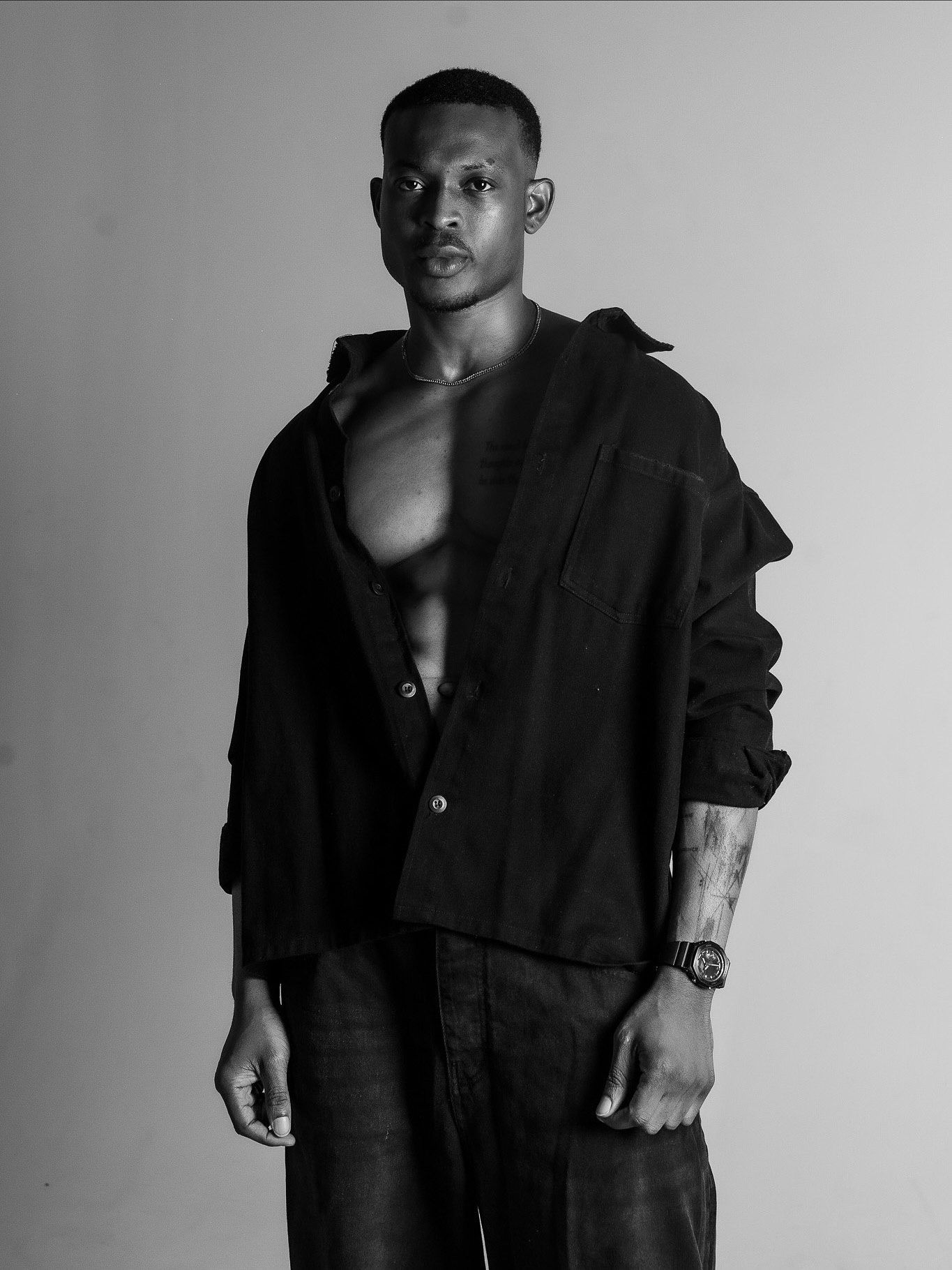Black and white portrait of a muscular man in an open black shirt and pants, posing confidently against a light background.