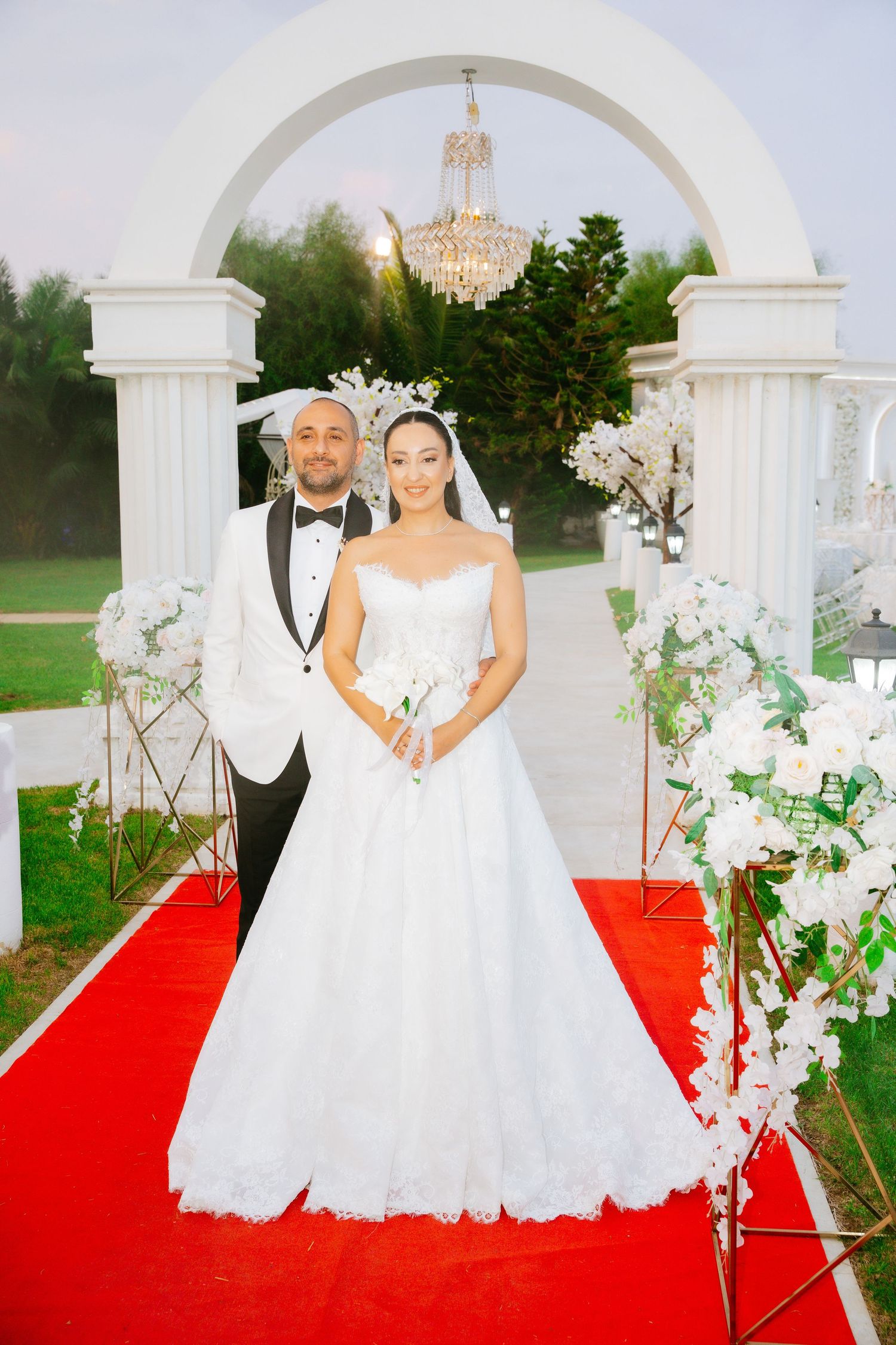 Bride and groom posing under white arch on red carpet at outdoor wedding ceremony with floral decorations.