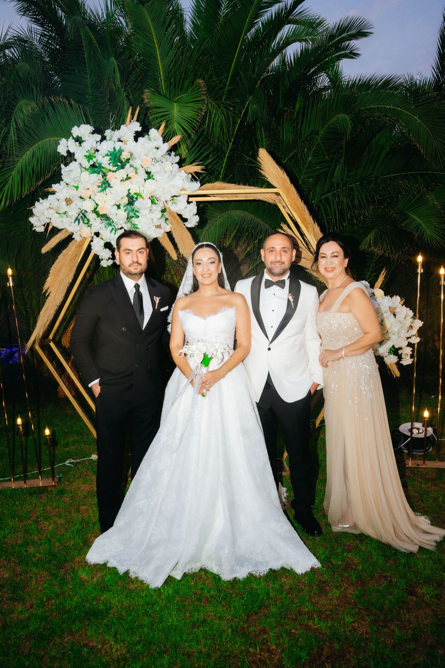 Wedding party of four posing before a geometric gold arch with white flowers and palm trees at an outdoor evening ceremony.