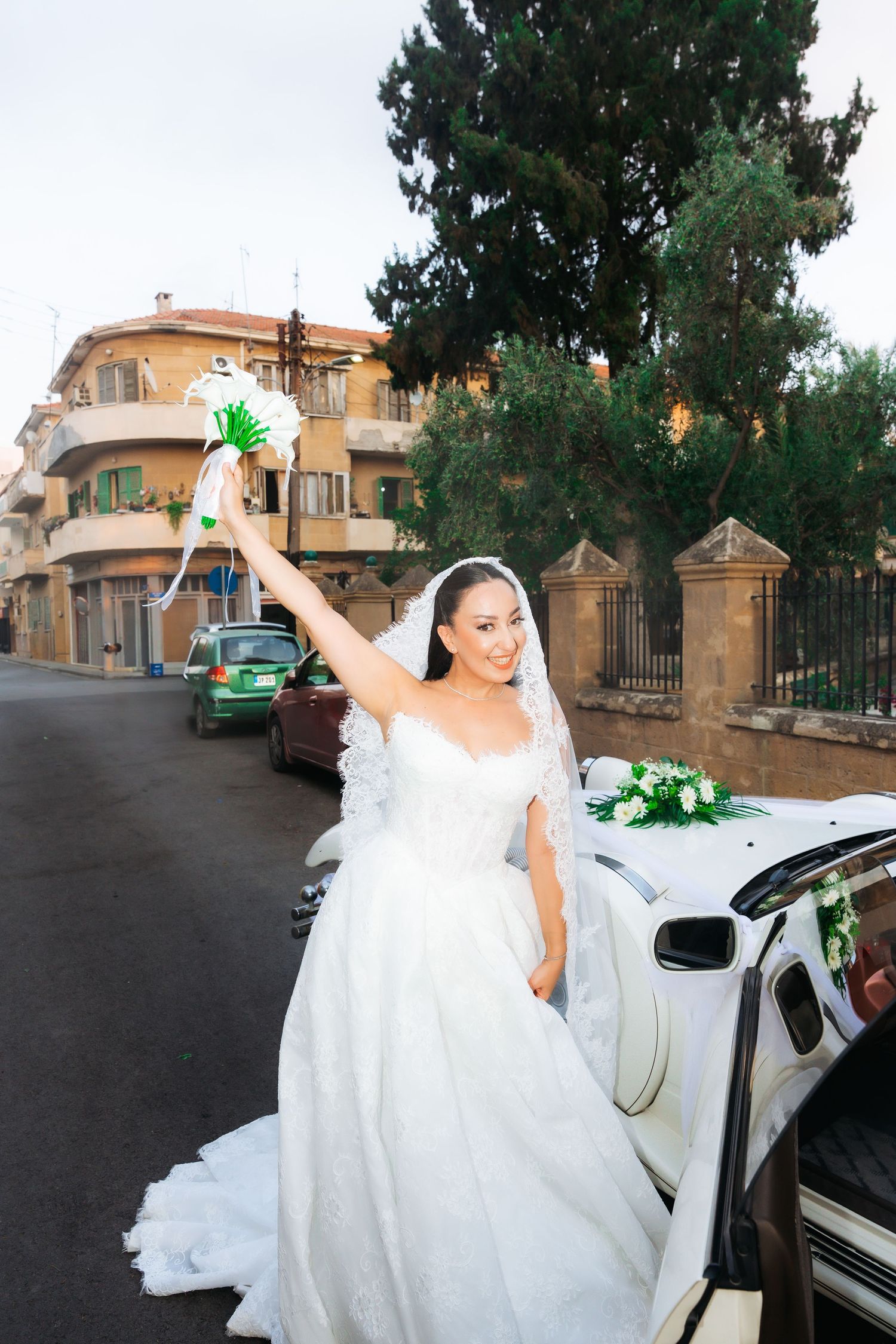Smiling bride in white gown and veil raises bouquet beside decorated white car on a tree-lined street.