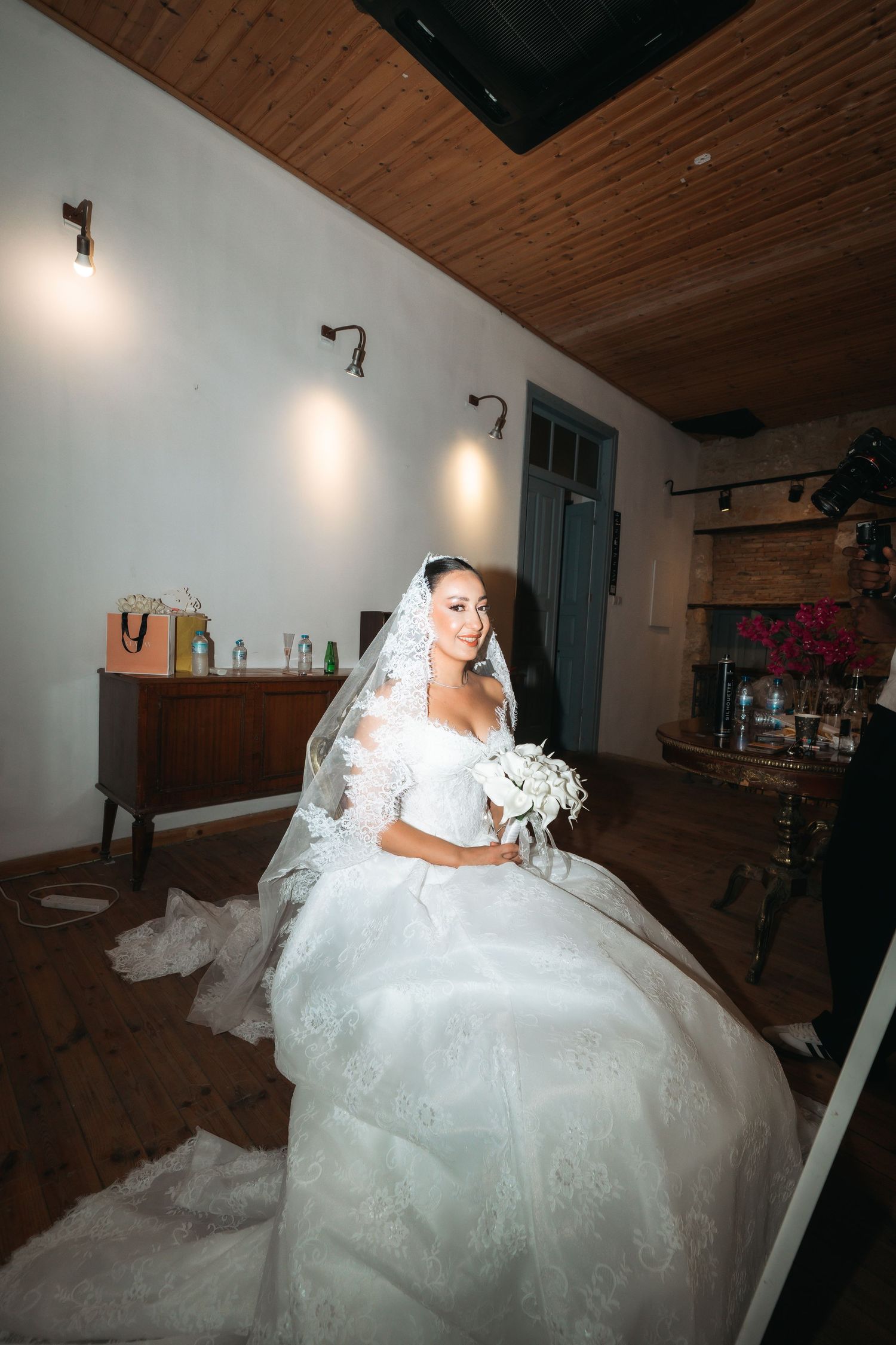 Elegant bride in white lace gown and veil holding bouquet, seated in warmly lit rustic indoor setting.