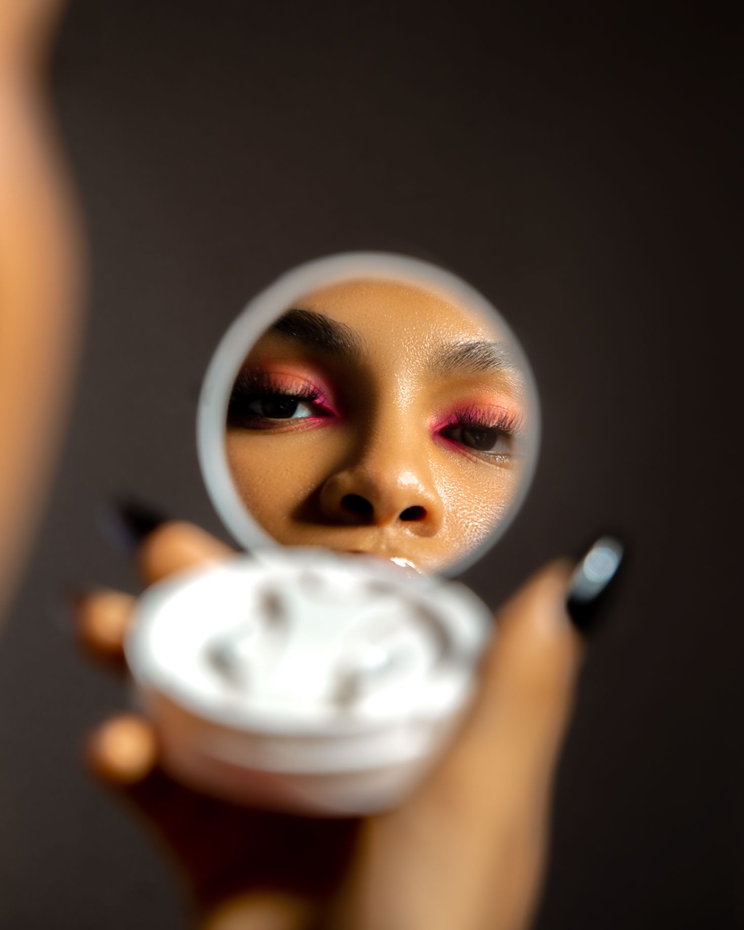 Woman with pink eyeshadow and black nails holds a compact mirror reflecting her bold makeup look.