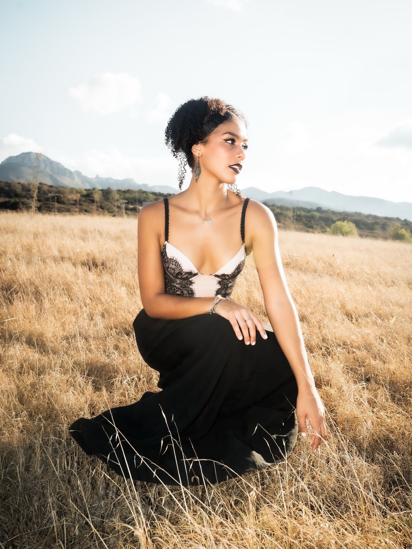 Woman in black skirt and lace top sitting in golden field with mountain backdrop.