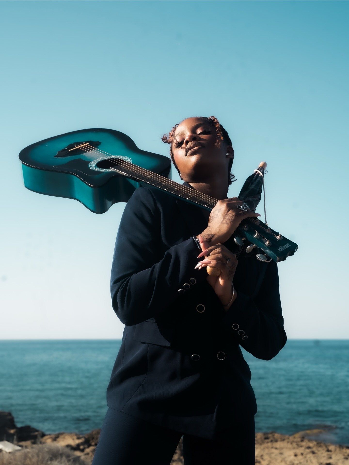 Musician in navy blazer holds teal acoustic guitar on shoulder against ocean horizon backdrop.