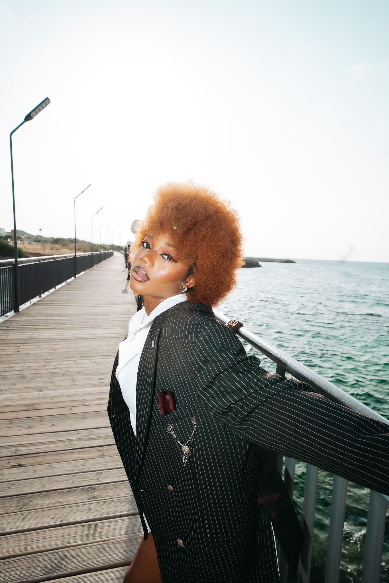 Woman with auburn afro in pinstripe blazer leans on railing of scenic ocean pier.
