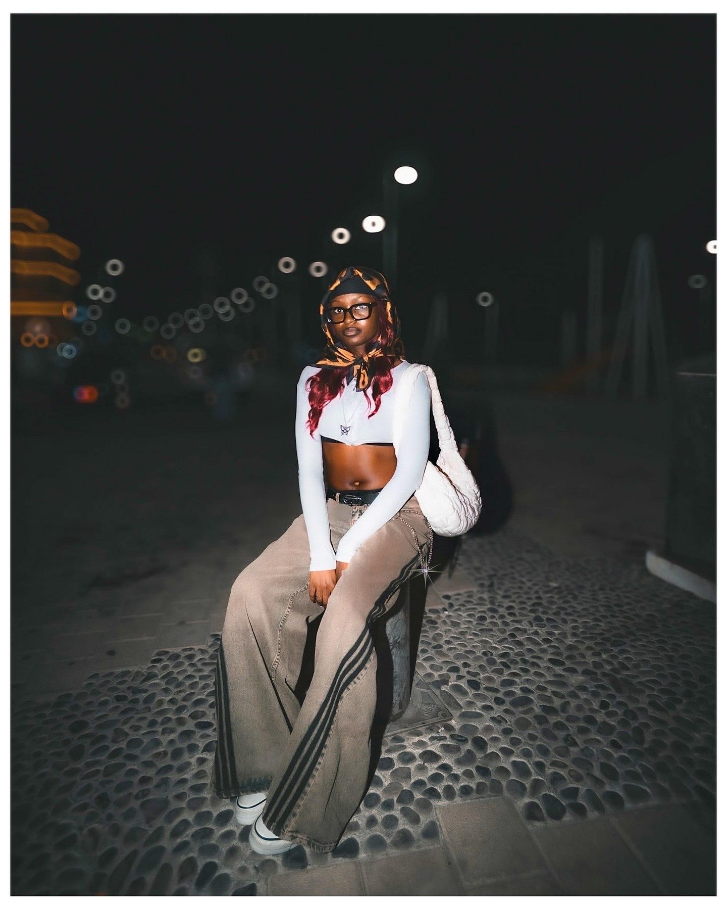 Woman in white crop top and wide-leg pants poses outdoors at night under bokeh lights.