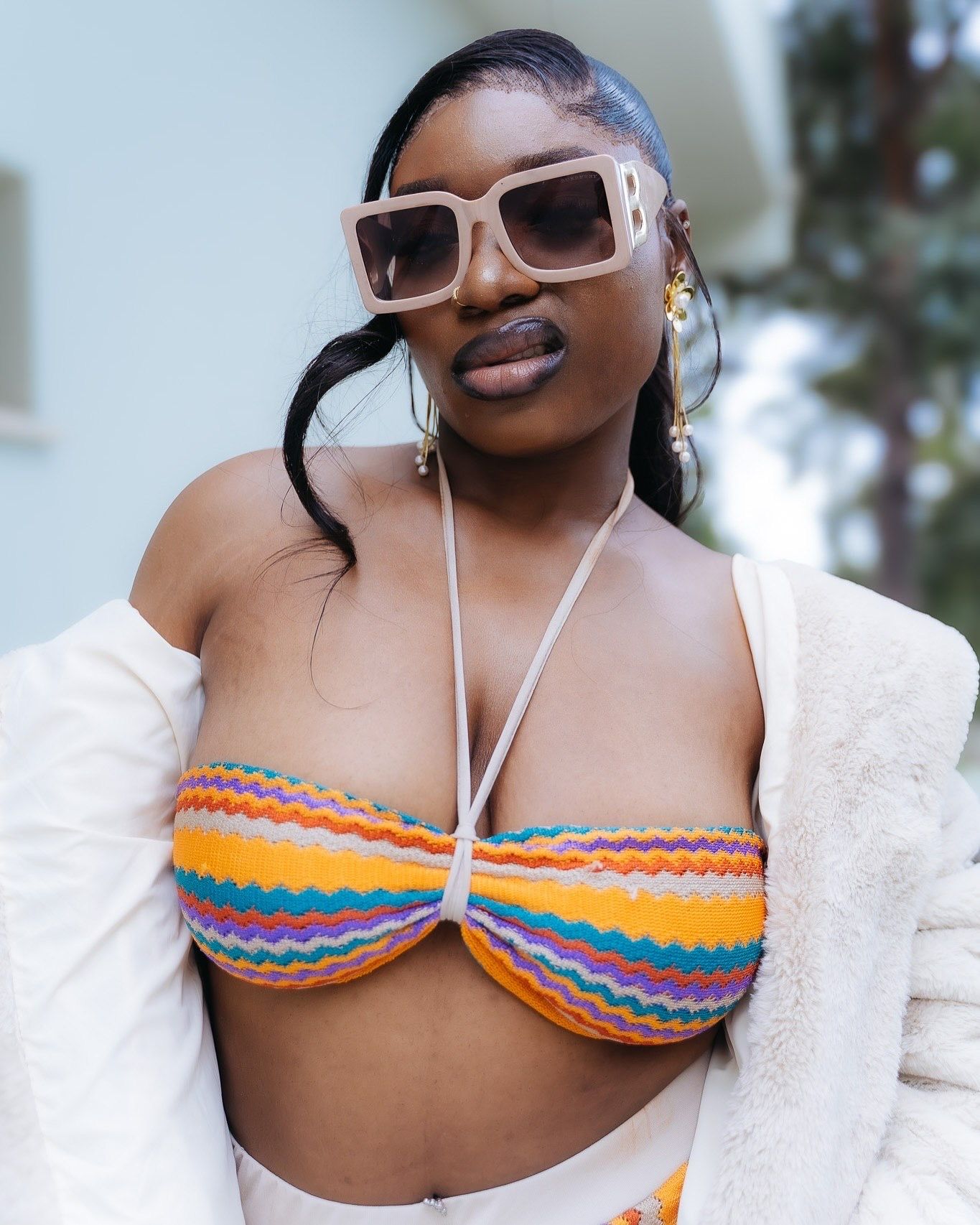 Woman wearing colorful zigzag bikini top, oversized sunglasses, gold earrings, and white jacket outdoors.