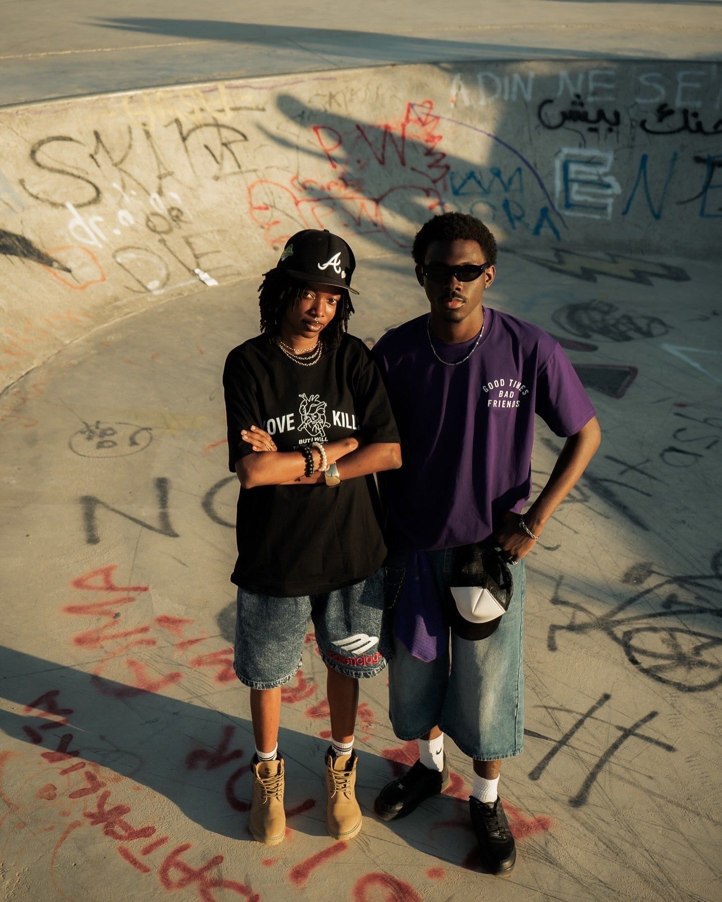 Two young men posing at a graffiti-covered skate park, wearing streetwear including denim shorts and graphic tees.