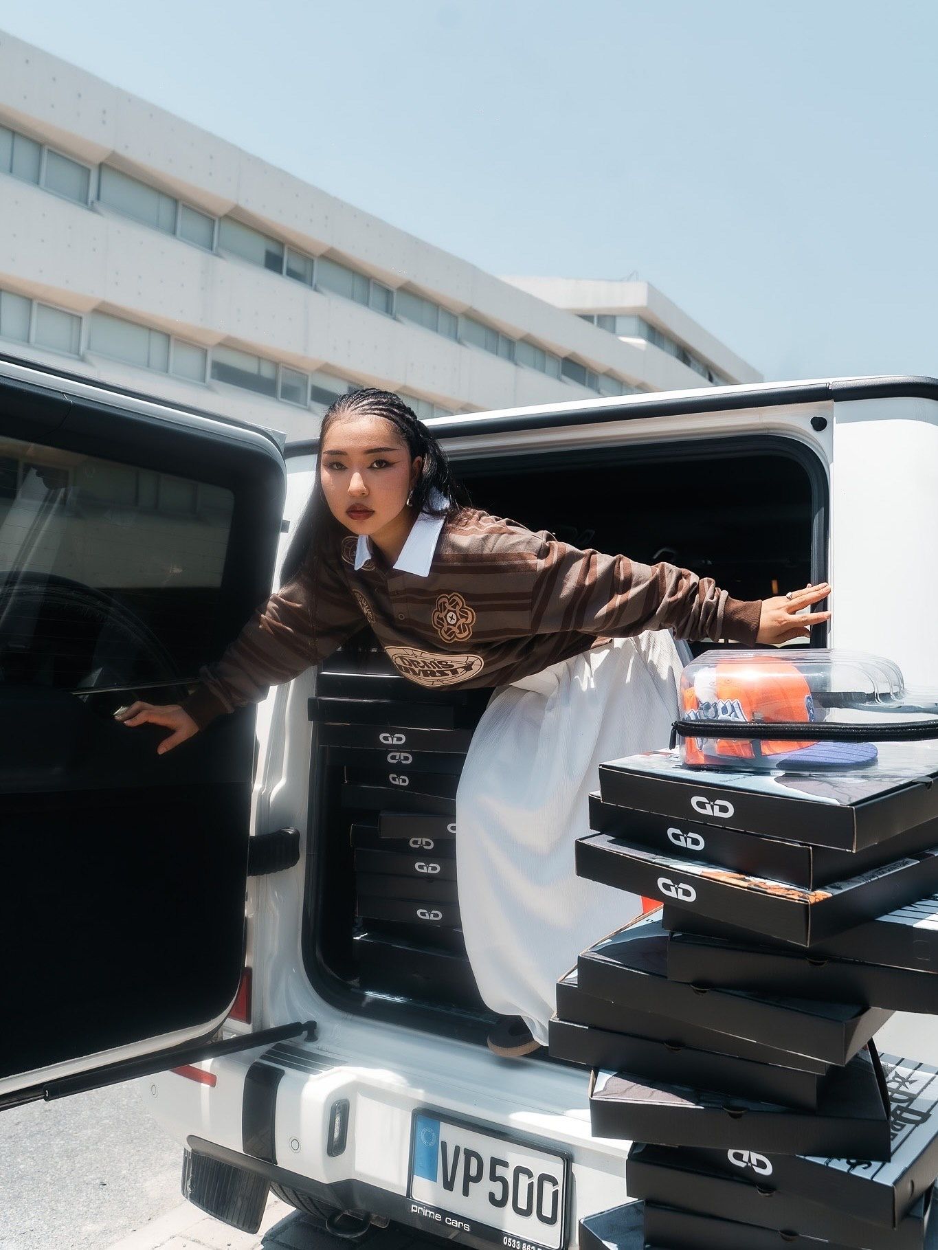 Woman in brown jacket leans out of white SUV trunk surrounded by stacked black boxes outdoors.
