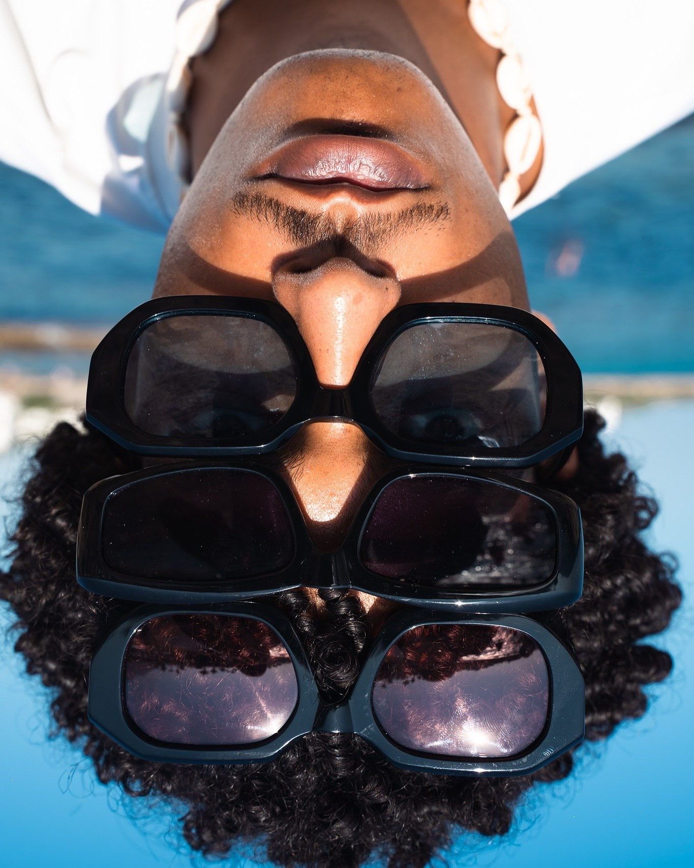 Person with curly hair wearing multiple stacked black sunglasses, photographed upside down against a blue background.