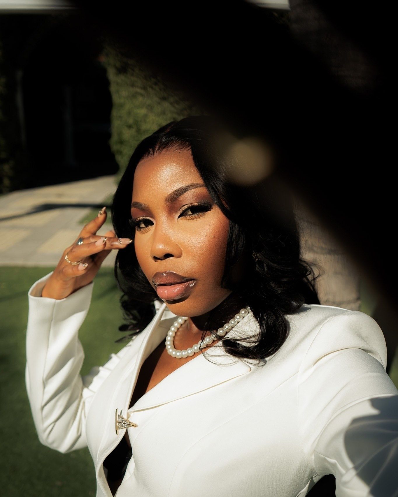 A stylish woman in a white blazer poses outdoors, wearing a pearl necklace and rings, with sleek black hair.